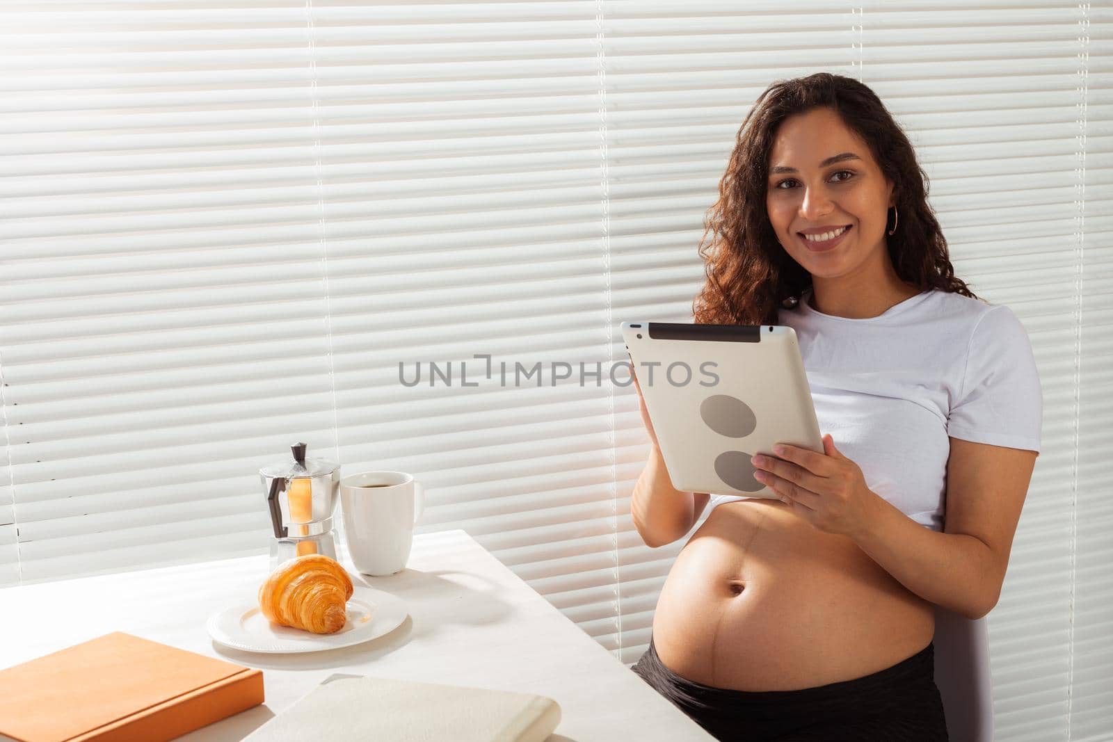 Hispanic pregnant woman using digital tablet while breakfast. Technology, pregnancy and maternity leave.
