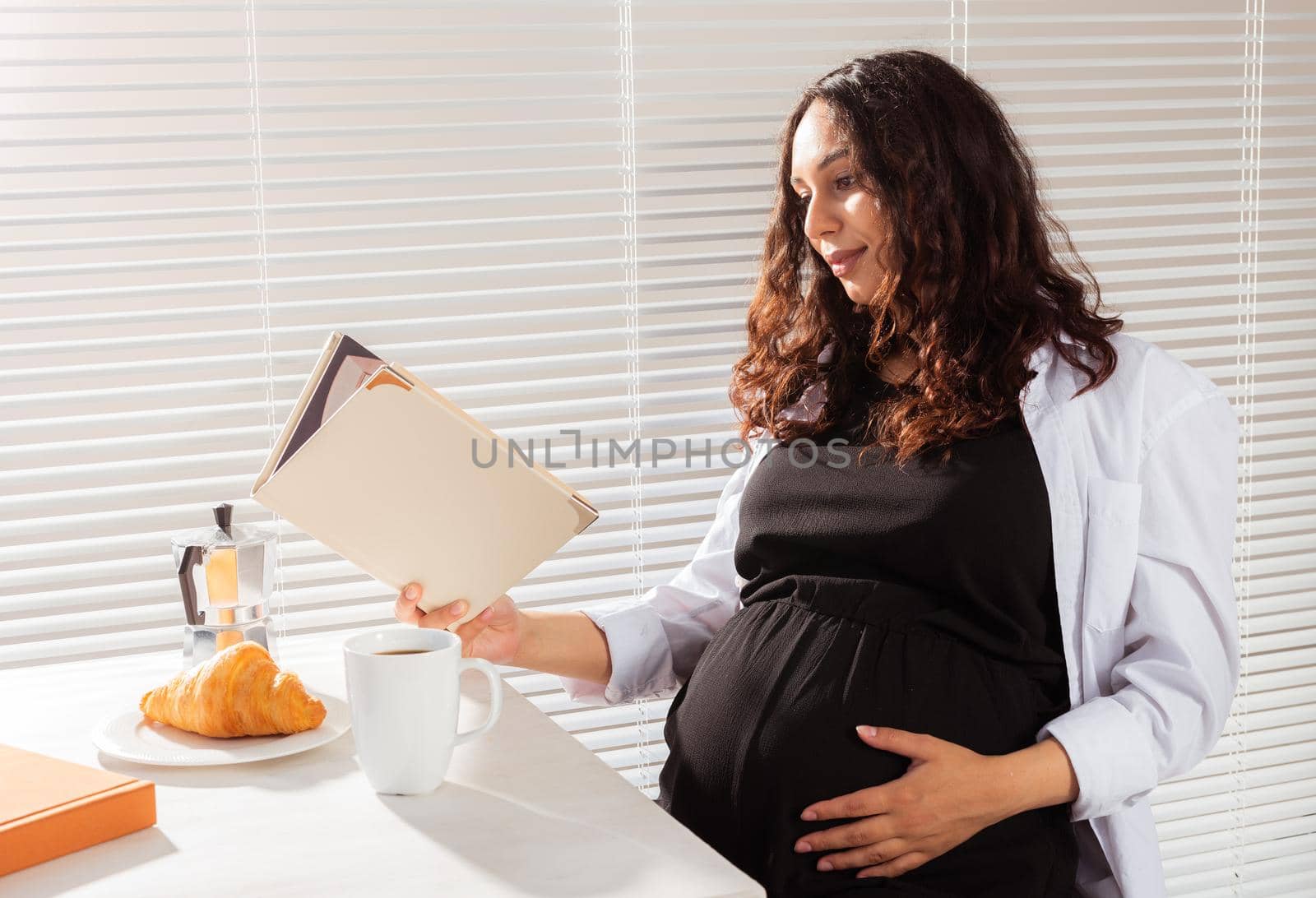 Positive young beautiful woman reads book during morning breakfast with coffee and croissants. Pleasant morning and expectation of meeting baby
