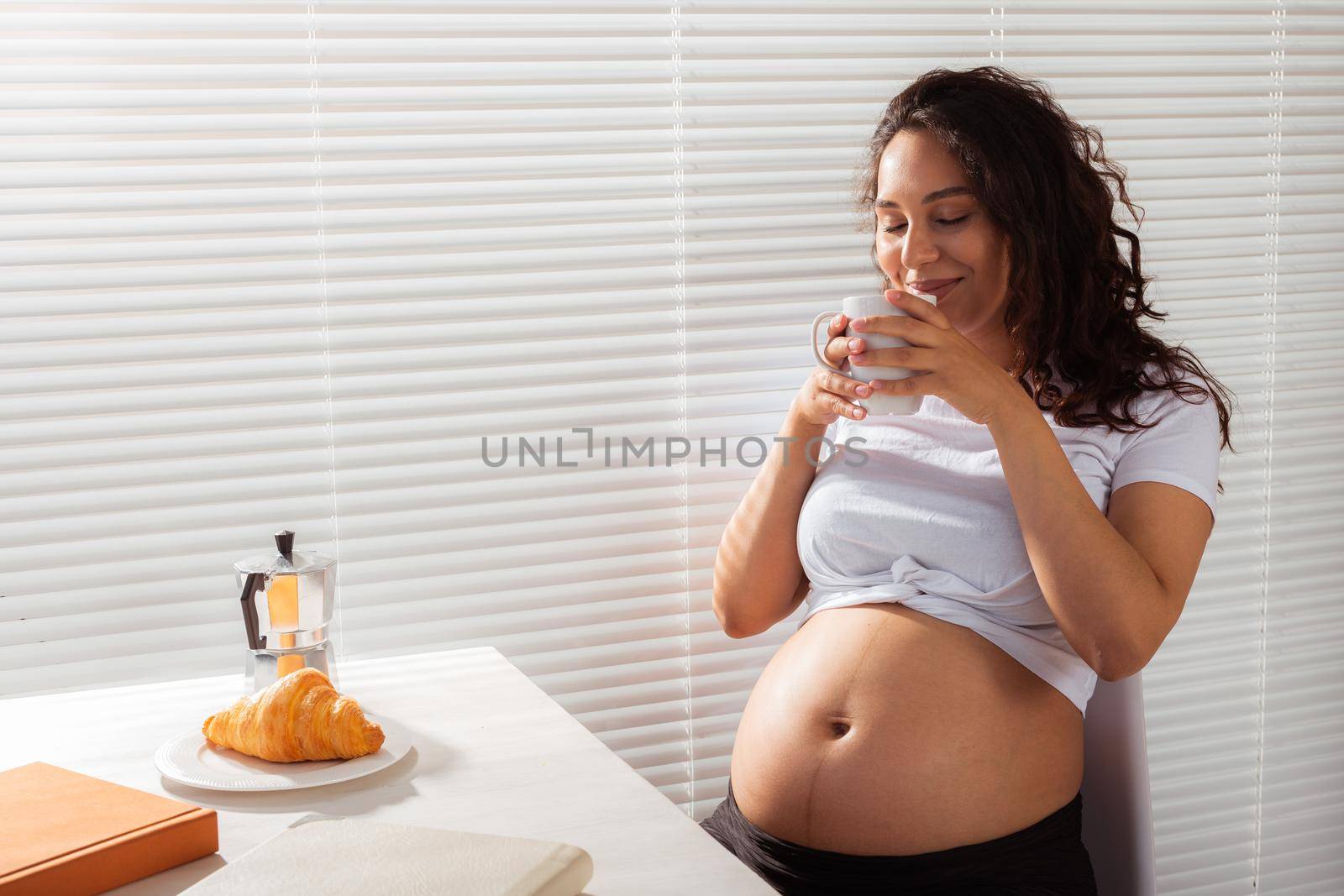Joyful pregnant young beautiful woman looks through blinds during her morning breakfast with coffee and croissants. Good morning and waiting for meeting with a baby