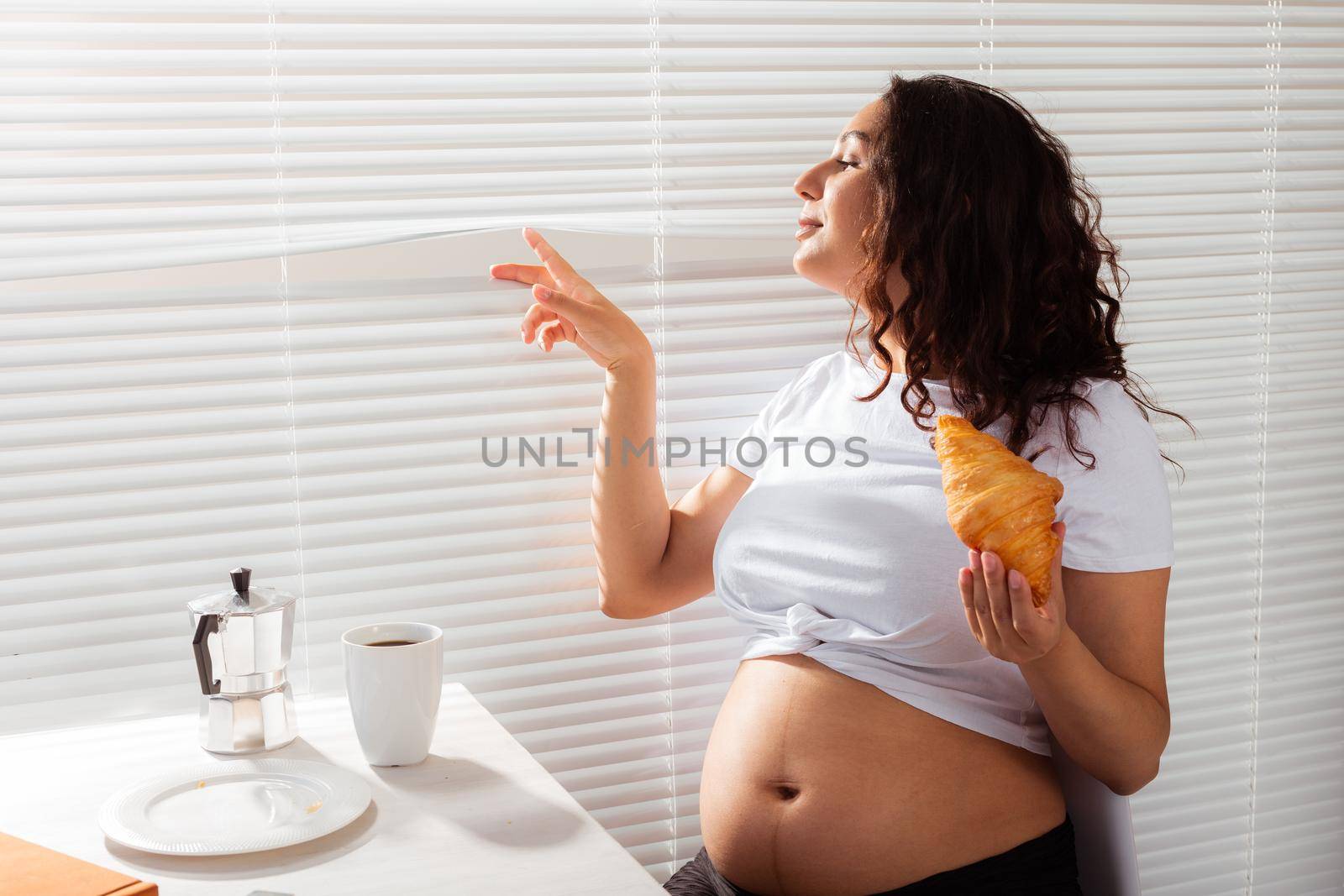 Happy pregnant young beautiful woman eating croissant and looking througt blinds during morning breakfast. Pleasant morning and positive attitude during pregnancy
