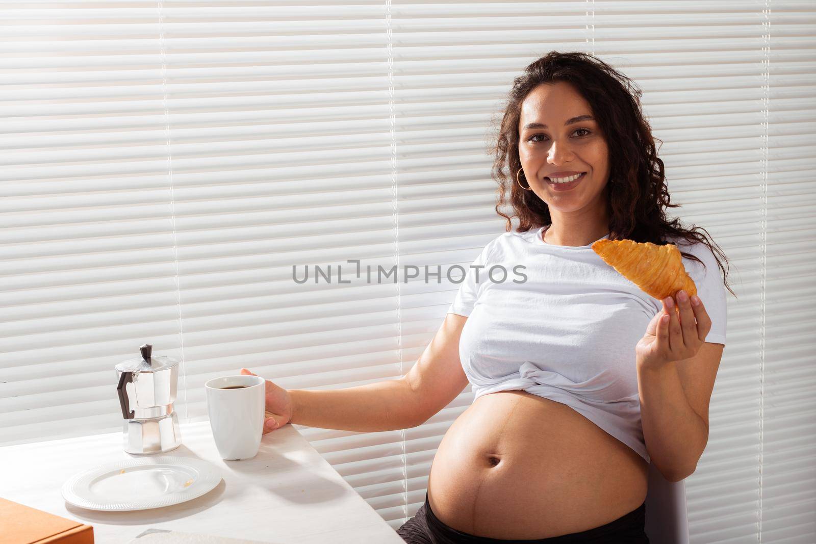 Joyful pregnant young beautiful woman eating croissant and drinking tea during morning breakfast. Concept of pleasant morning and expectation of meeting baby.