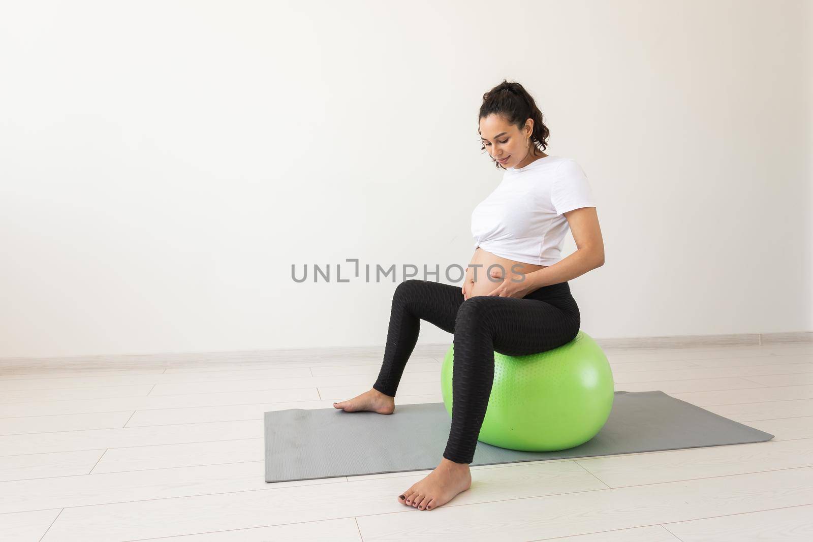 A young pregnant woman doing relaxation exercise using a fitness ball while sitting on a mat. Copyspace. by Satura86