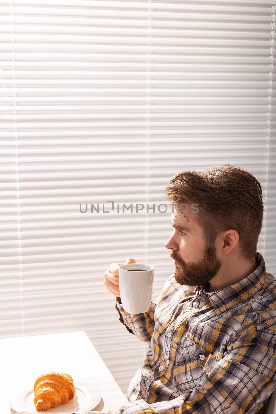 Pensive young bearded male businessman drinking cup of coffee on background of blinds. Concept of pleasant morning or lunch break. Copyspace
