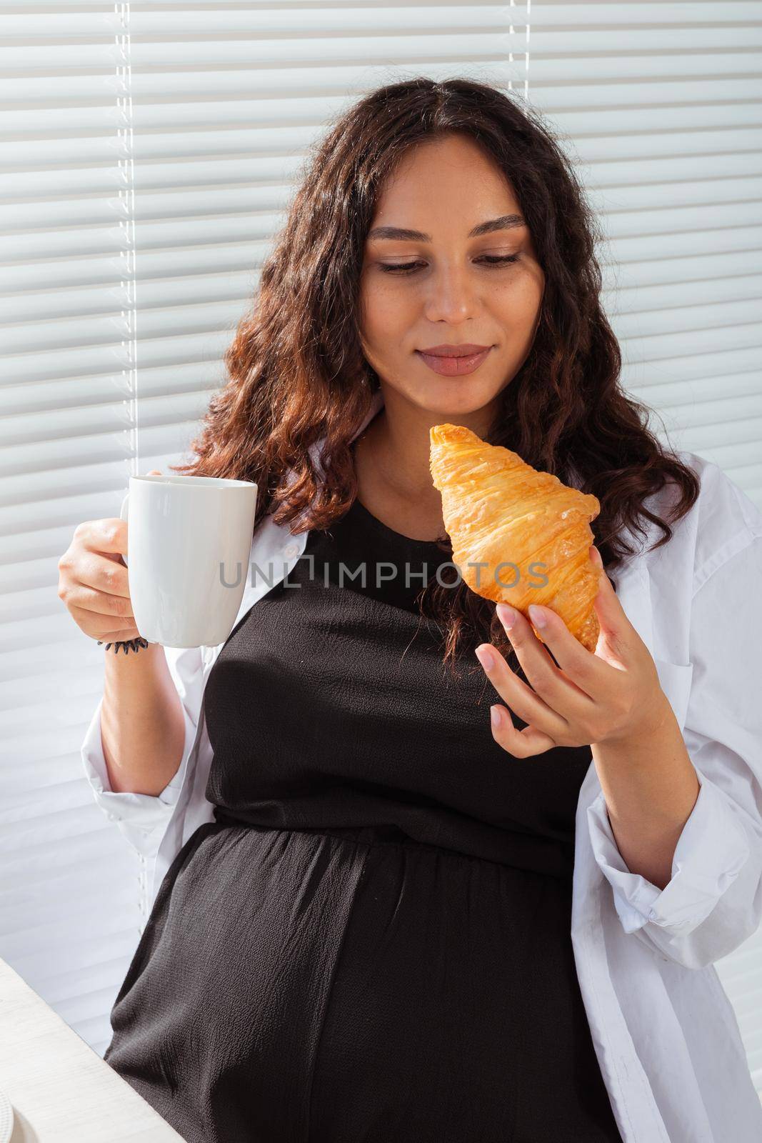Happy pregnant young beautiful woman eating croissant and looking througt blinds during morning breakfast. Pleasant morning and positive attitude during pregnancy