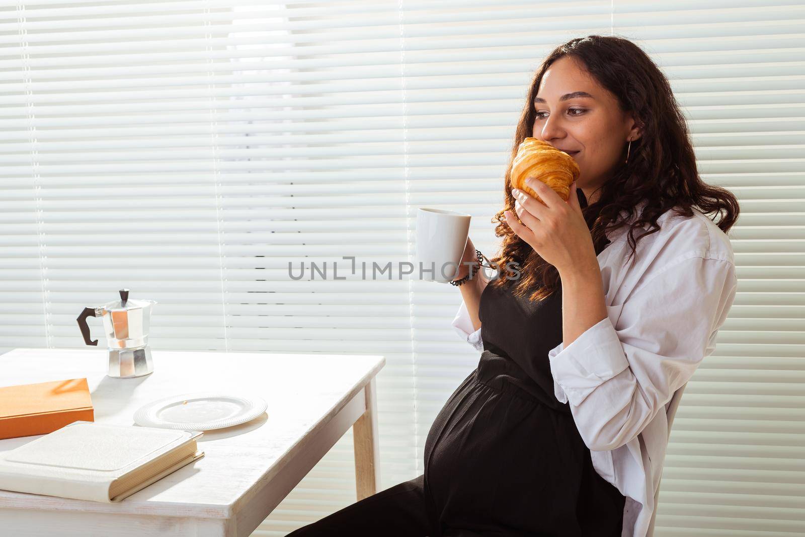 Happy pregnant young beautiful woman eating croissant and looking througt blinds during morning breakfast. Pleasant morning and positive attitude during pregnancy