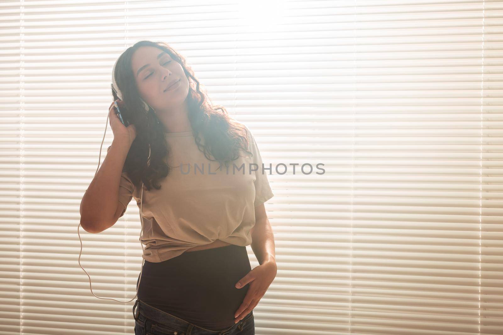 Curly-haired brunette pacified pregnant woman listens to pleasant classical music using smartphone and headphones. Concept of a soothing mood before meeting baby