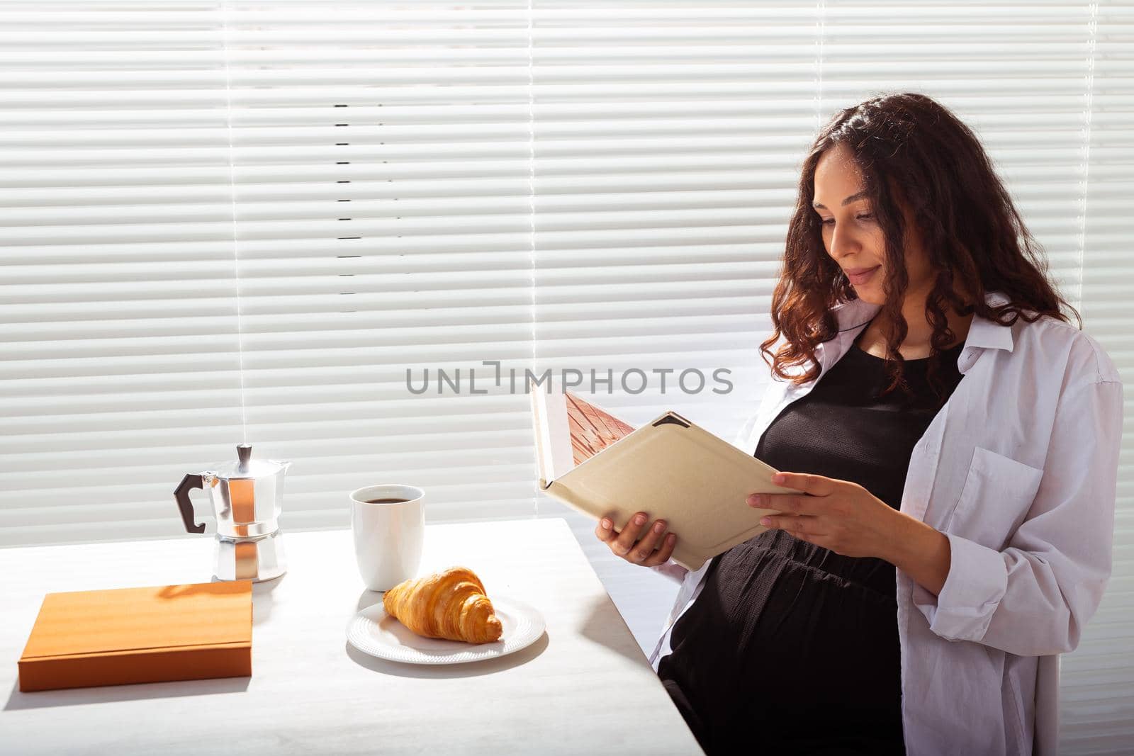 Side view of happy young beautiful woman reading book while having morning breakfast with coffee and croissants on background of blinds. Good morning and pleasant lunch break