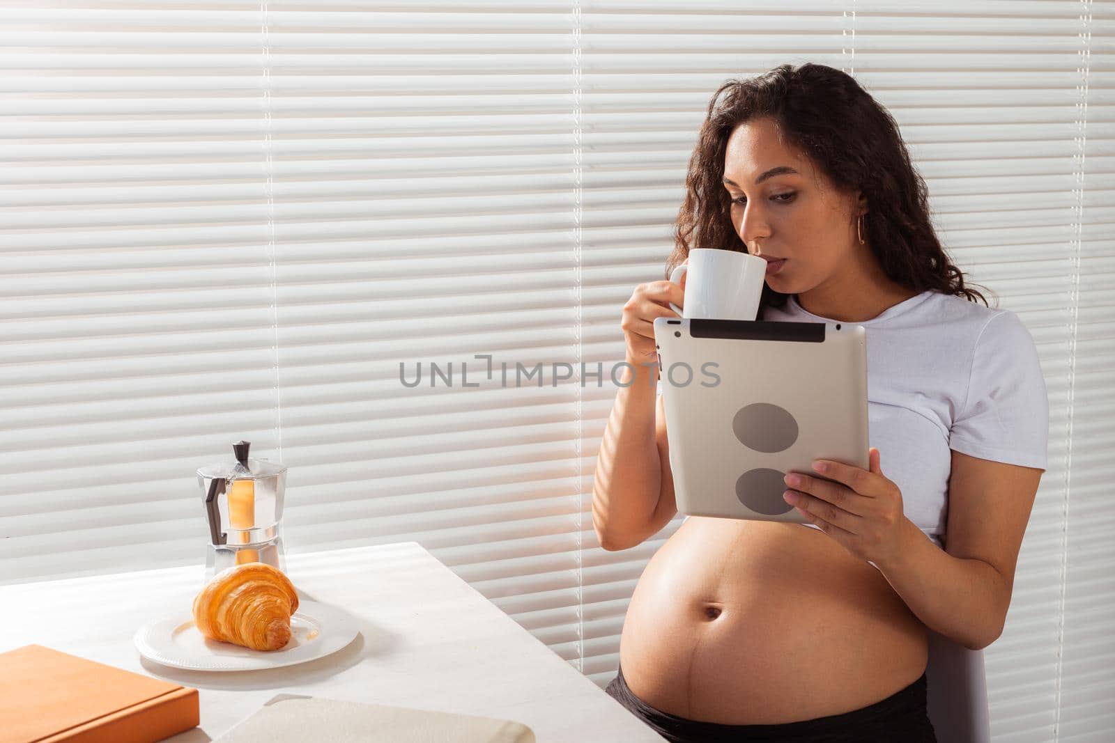 Hispanic pregnant woman using digital tablet while breakfast. Technology, pregnancy and maternity leave.