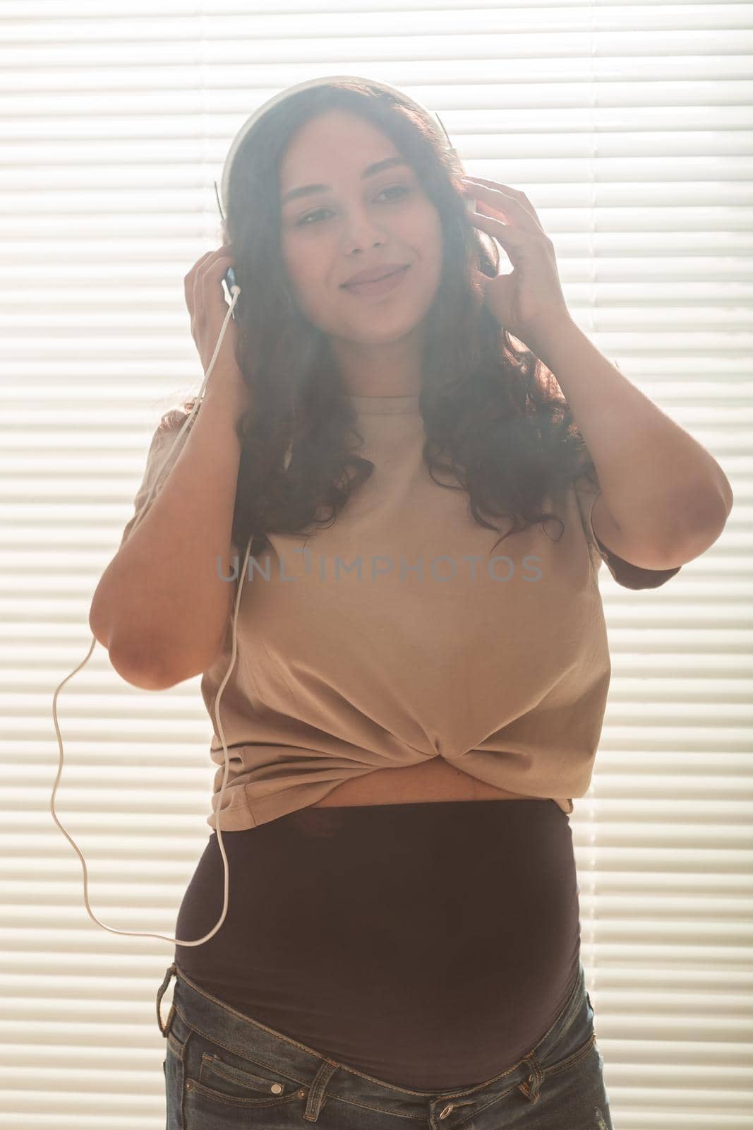 Curly-haired brunette pacified pregnant woman listens to pleasant classical music using smartphone and headphones. Concept of a soothing mood before meeting baby