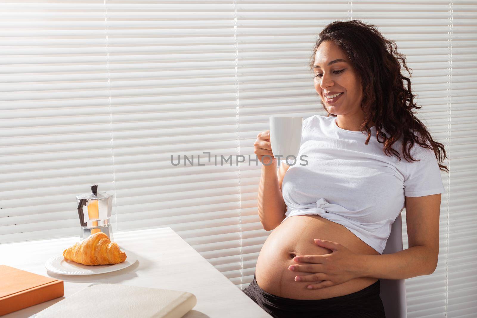 Joyful pregnant young beautiful woman looks through blinds during her morning breakfast with coffee and croissants. Good morning and waiting for meeting with a baby