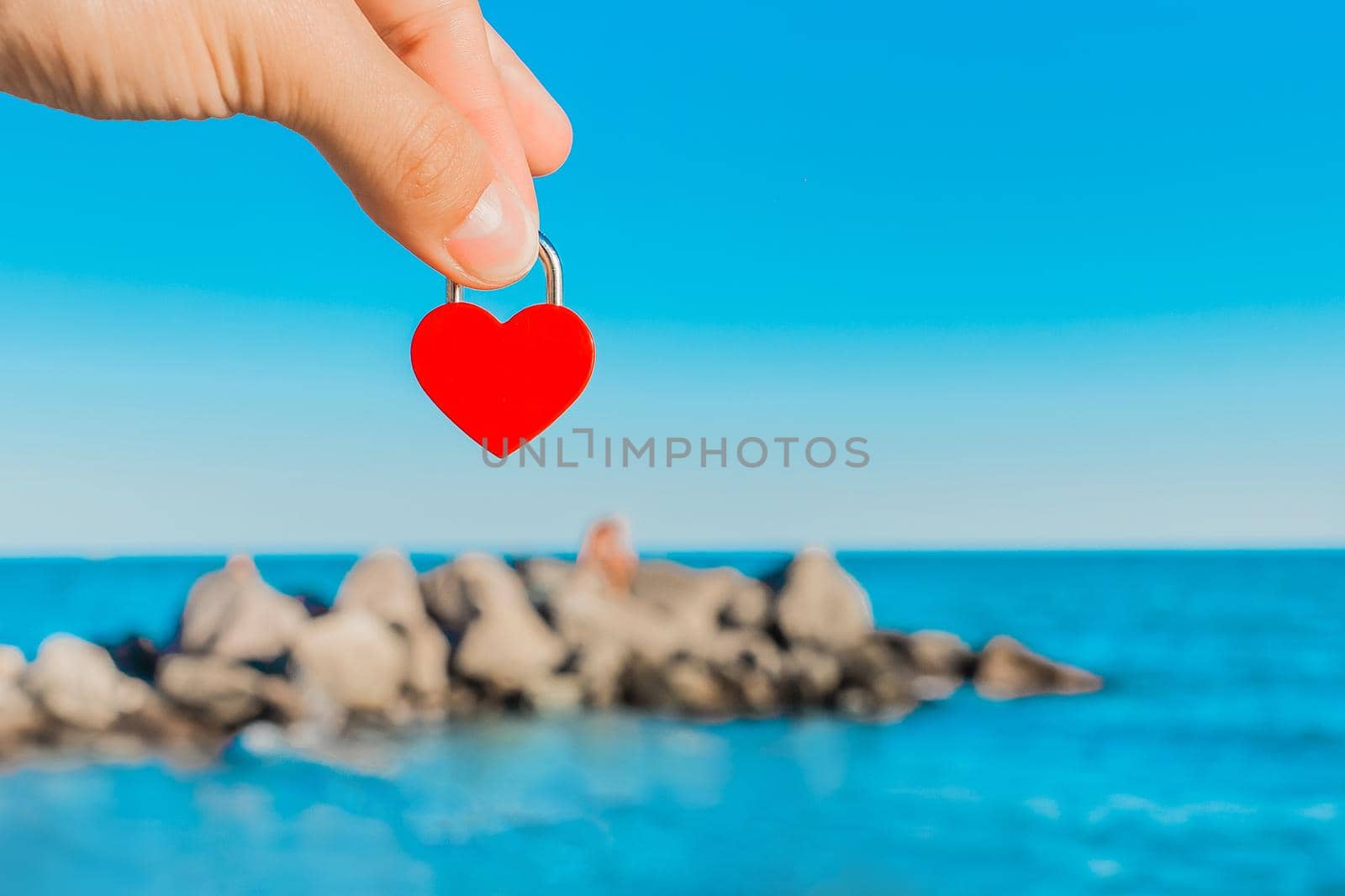The man's hand holds a small red heart in the form of a castle against the backdrop of the sea coast and the stones of the breakwater on the horizon line. Symbol or sign of love.