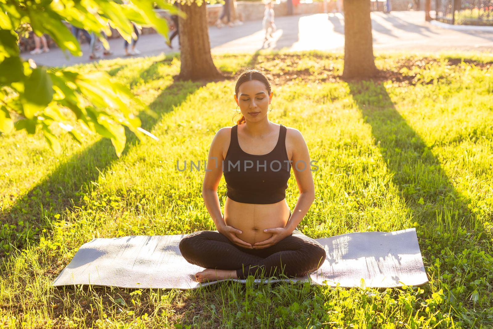 Cute young caucasian pregnant woman is meditating while sitting on a rug on the lawn on a sunny summer evening. Concept of pacification