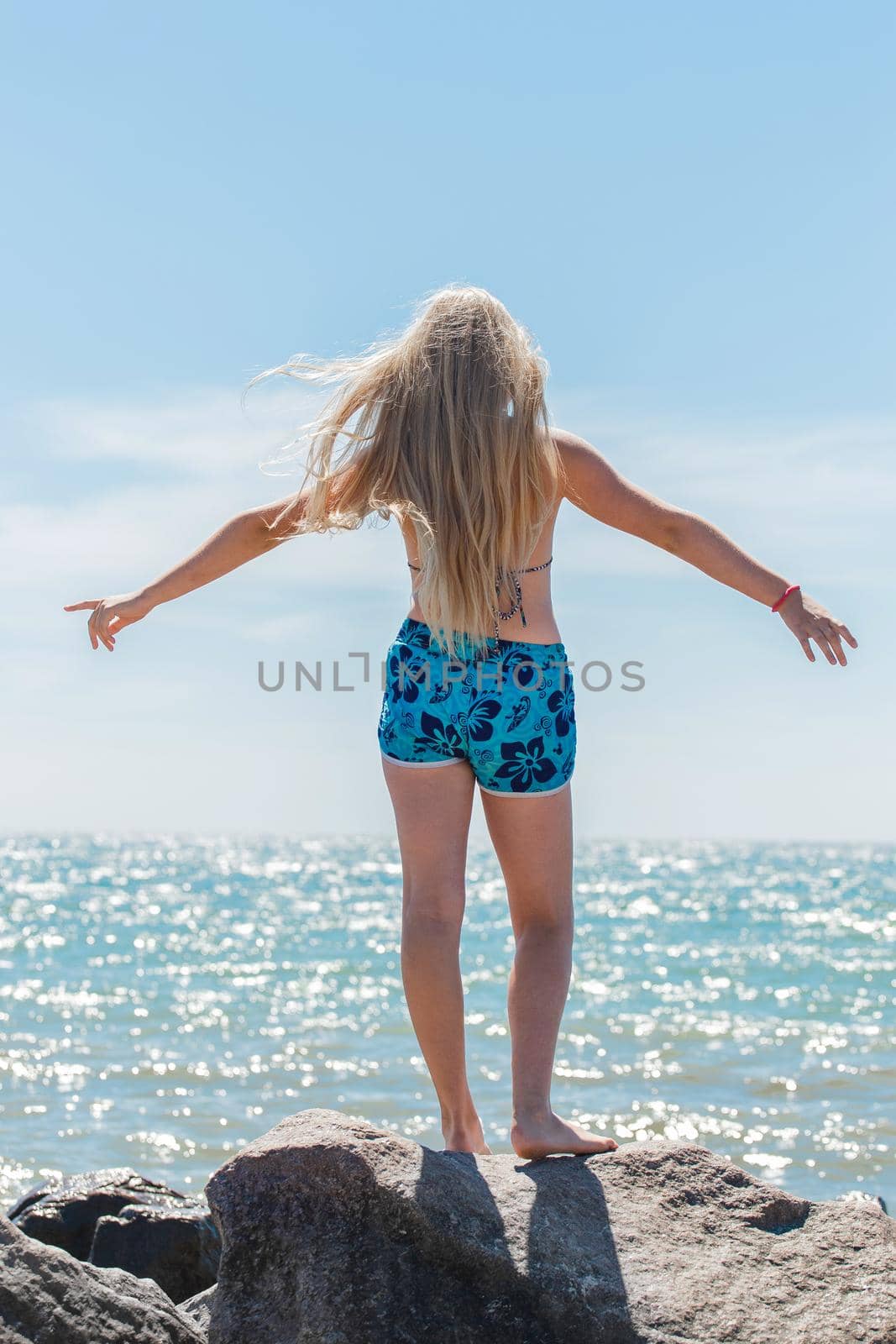 A young blonde girl, a teenager of European appearance in a swimsuit and shorts poses spreading her hands to the sides on seashore against the blue sky and sea water by AYDO8