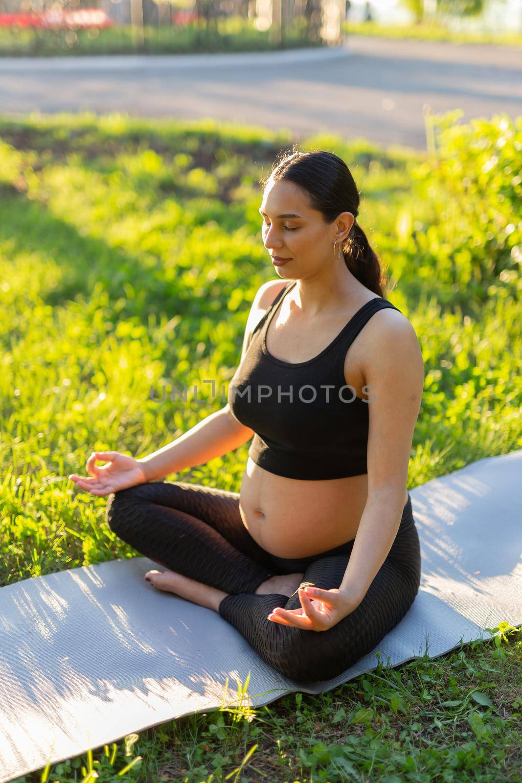 Peaceful young positive pregnant woman in gymnastic suit does yoga and meditate sitting on mat on green grass on sunny warm summer day.