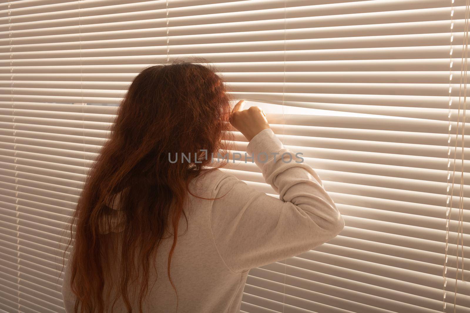 Rear view of beautiful young woman with long hair peeks through hole in the window blinds and looks out the window. Surveillance and curiosity