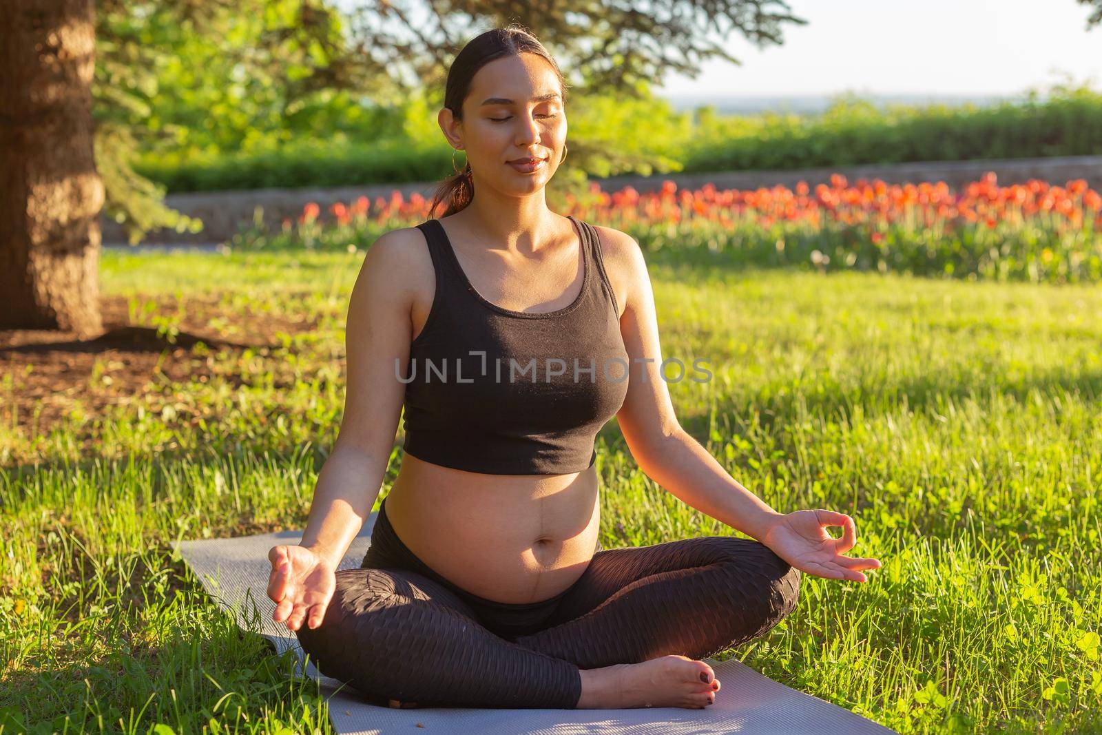 Cute young caucasian pregnant woman is meditating while sitting on a rug on the lawn on a sunny summer evening. Concept of pacification