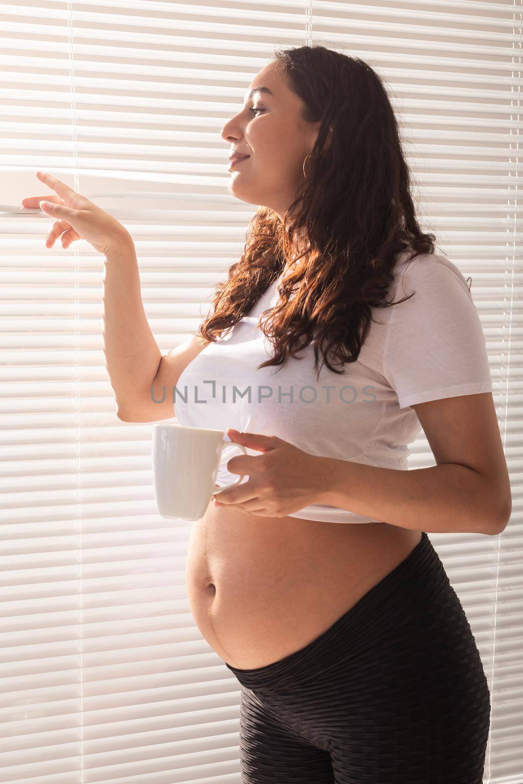 Curious young beautiful pregnant woman drinking tea and looking through the blinds at the window. Joy and good news while waiting for baby