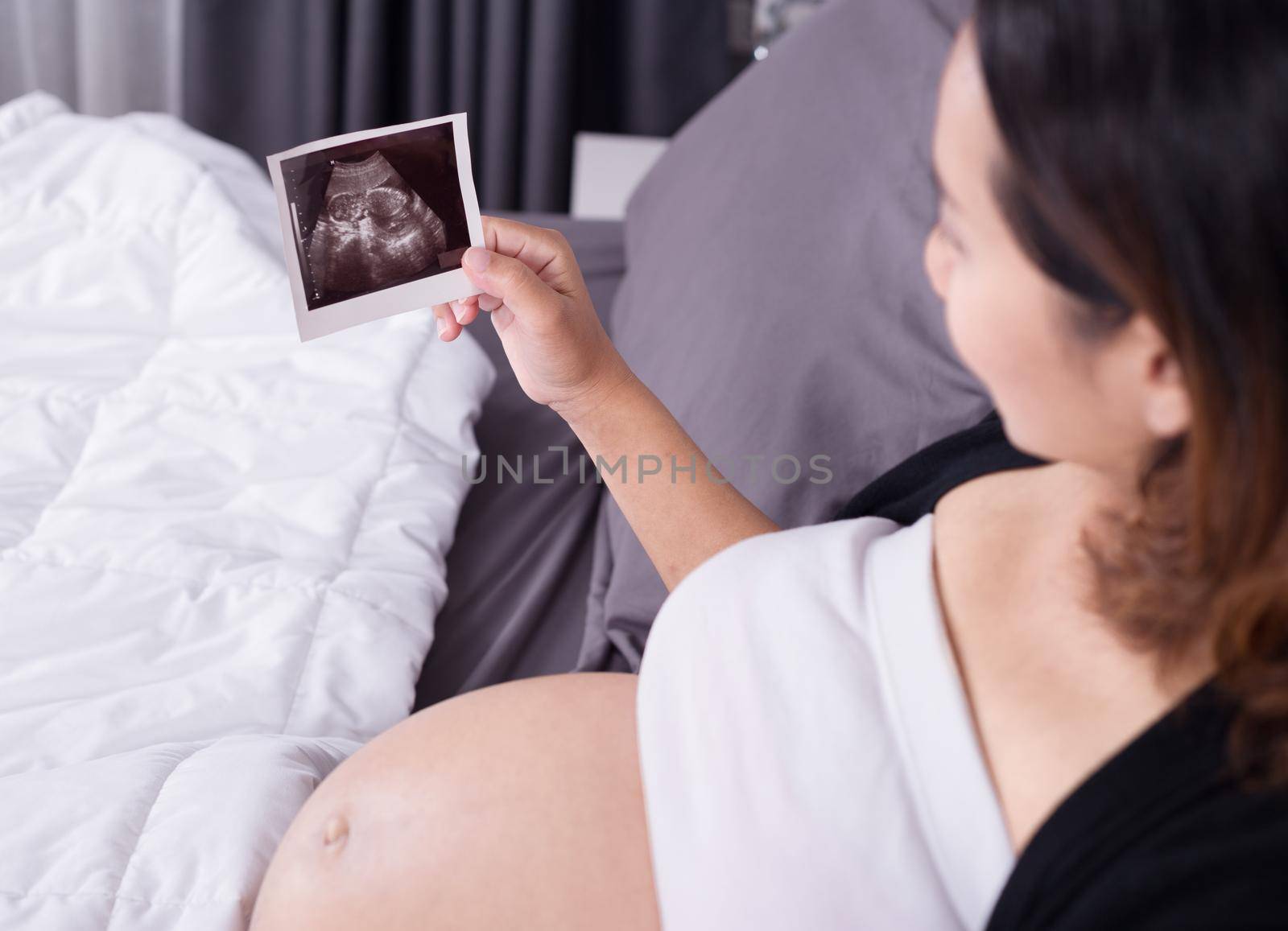 pregnant woman with ultrasound image of baby sitting on bed in bedroom at home by geargodz