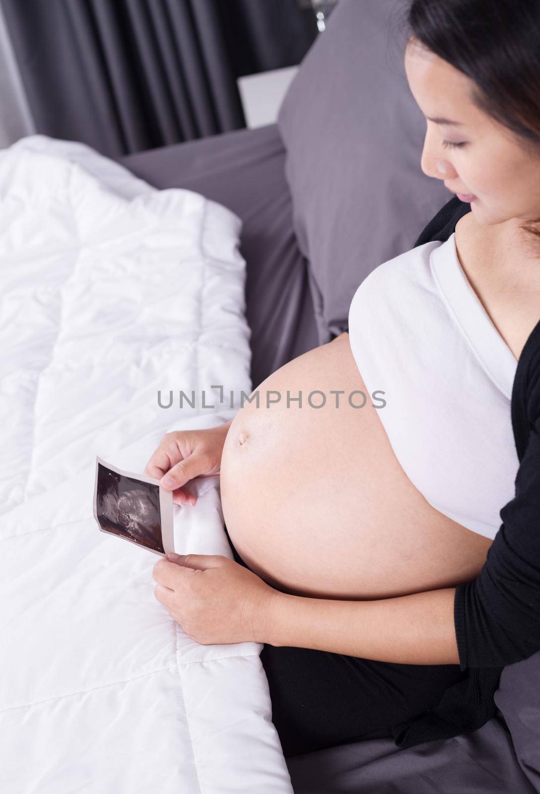 pregnant woman with ultrasound image of baby sitting on bed in bedroom at home by geargodz
