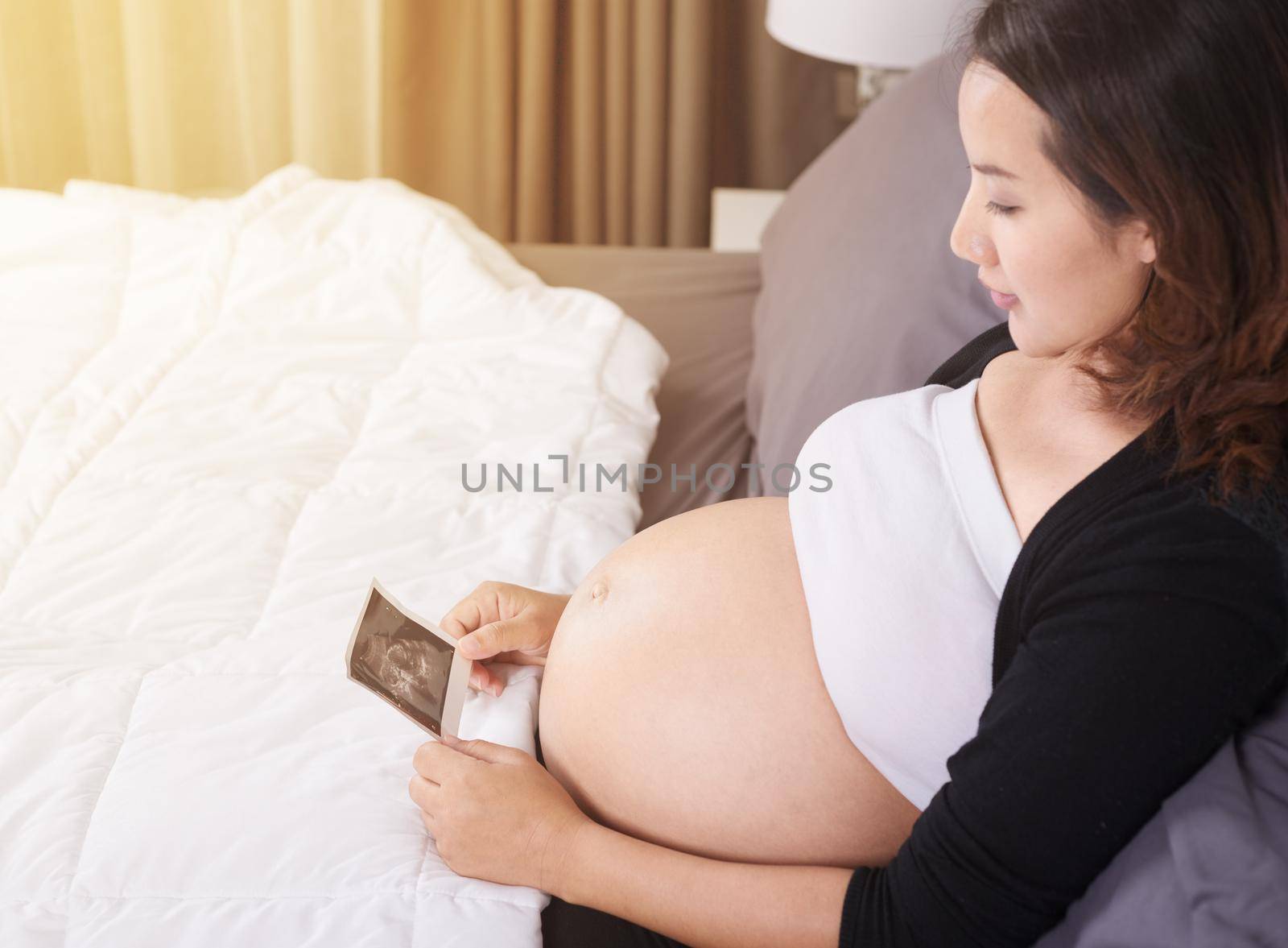 pregnant woman with ultrasound image of baby sitting on bed in the bedroom at home