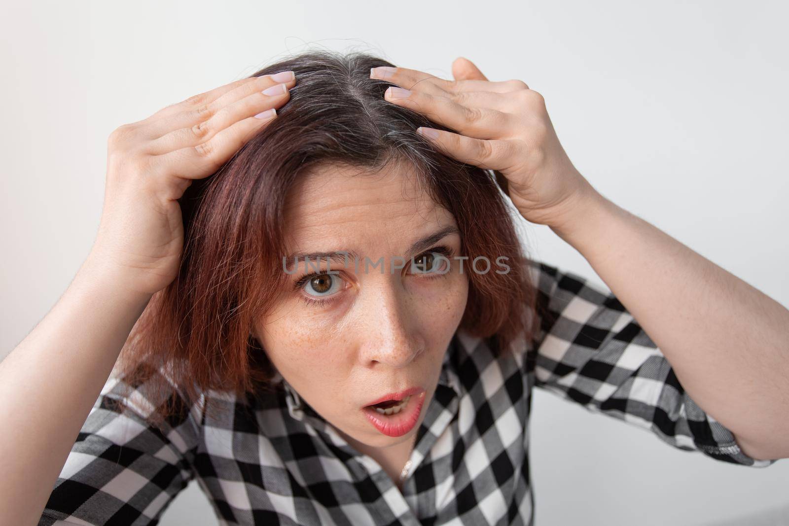 Horrified young woman looking in the bathroom mirror staring open mouthed at the first grey hair on her scalp, a first sign of ageing, or noticing that she is suffering from dandruff