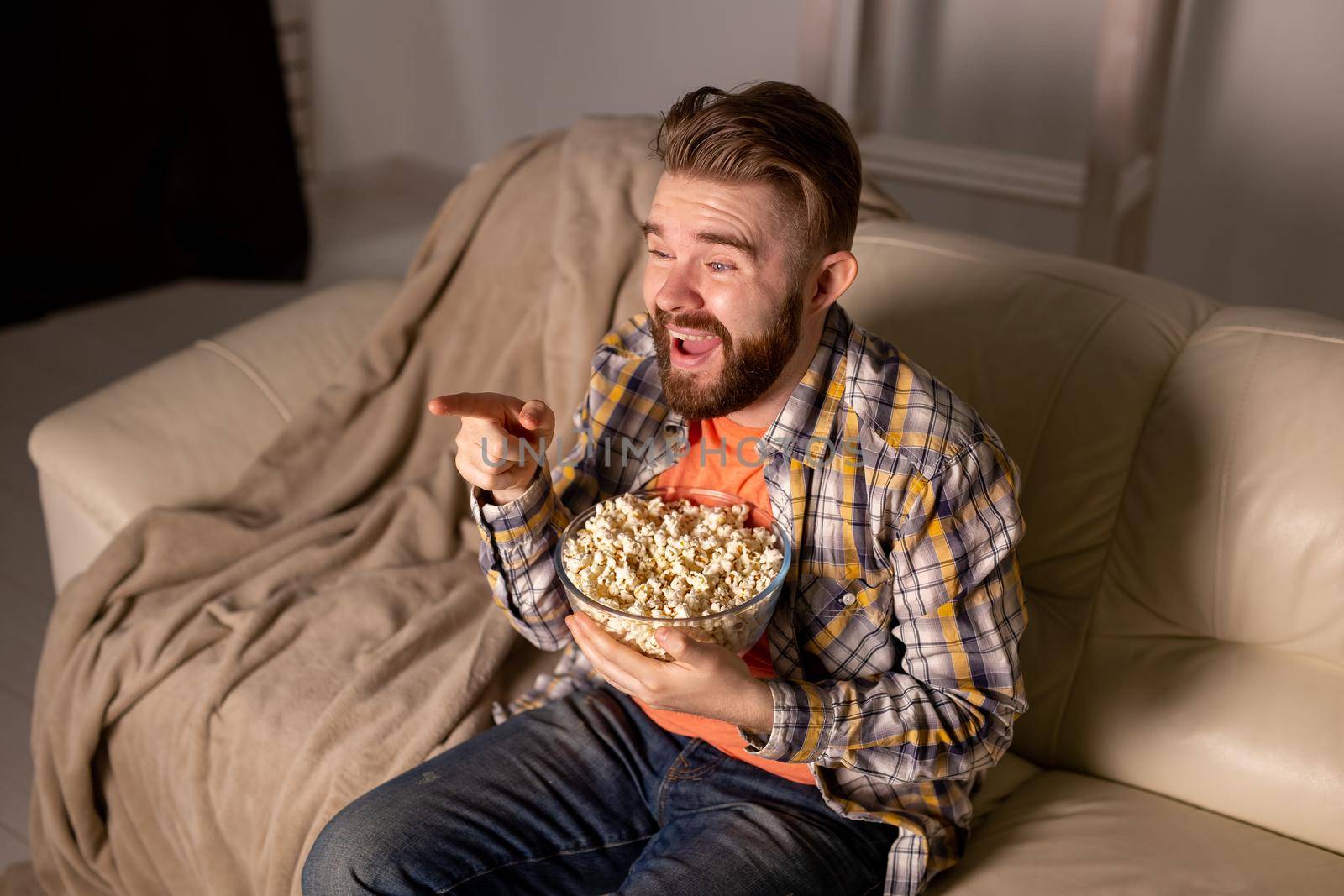 Portrait young man sitting on couch and watching TV holding remote and eats popcorn