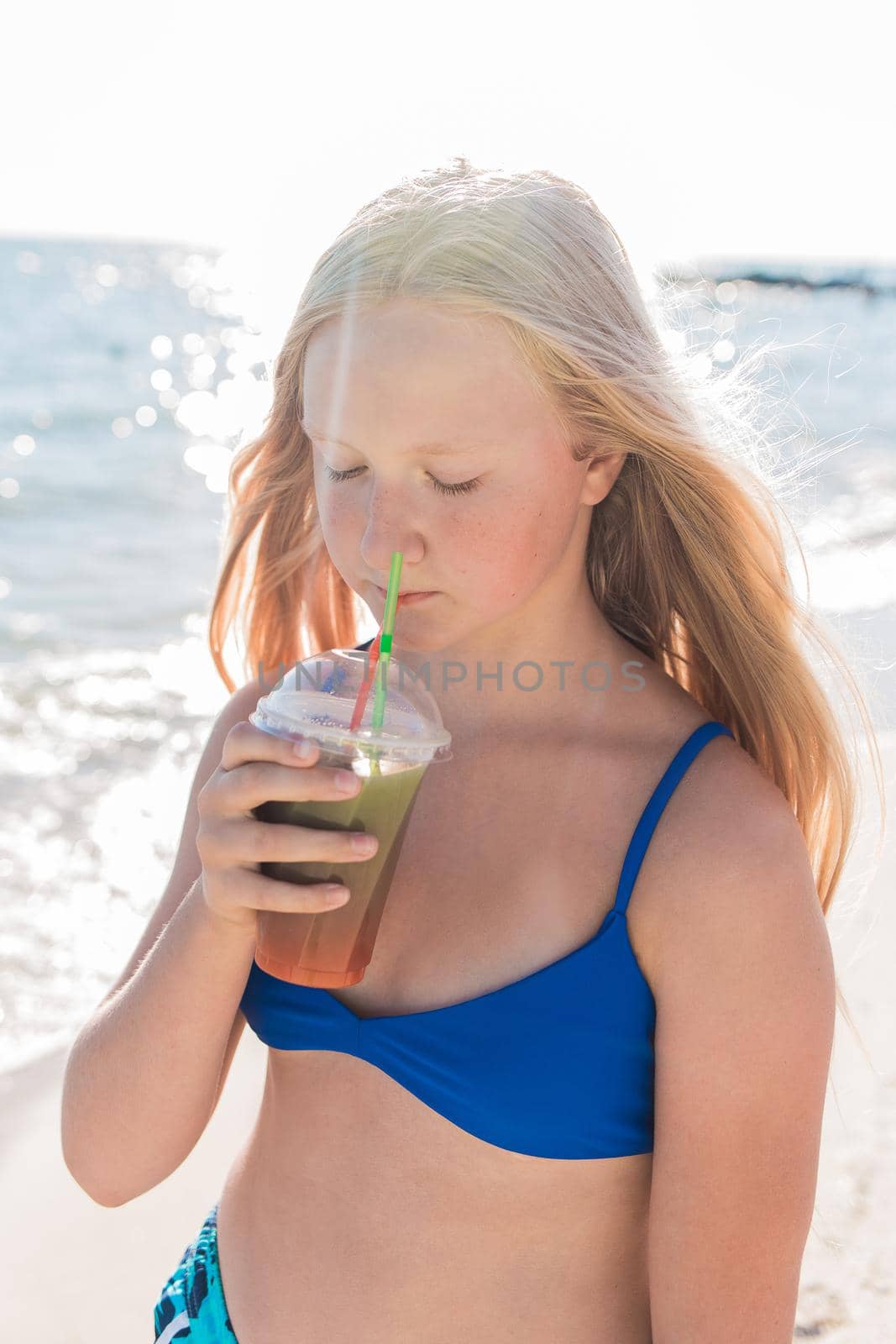 A young girl with blonde hair of European appearance, a teenager holds and drink a colored cold non-alcoholic cocktail in her hand against the background of the sea beach by AYDO8