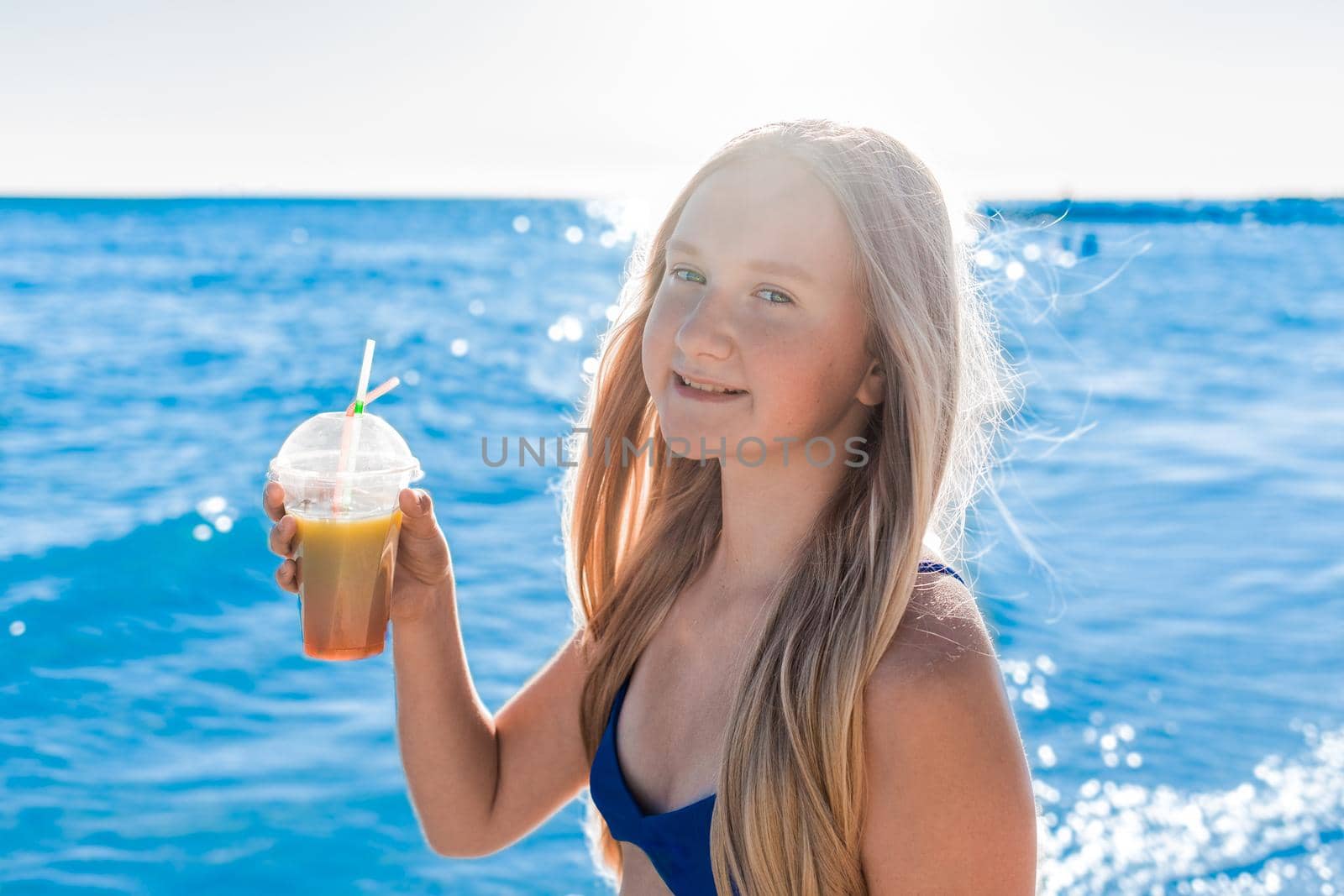 Young, cheerful attractive girl blonde of European appearance teenager in a blue swimsuit with a colored non-alcoholic cocktail in his hand against the background of the sea beach and the seashore by AYDO8