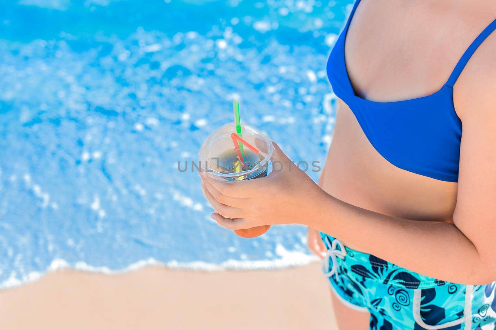 The hand of a young girl in a blue swimsuit and shorts holds a cooling, colored non-alcoholic cocktail against the background of the water of the sea beach and the shore.