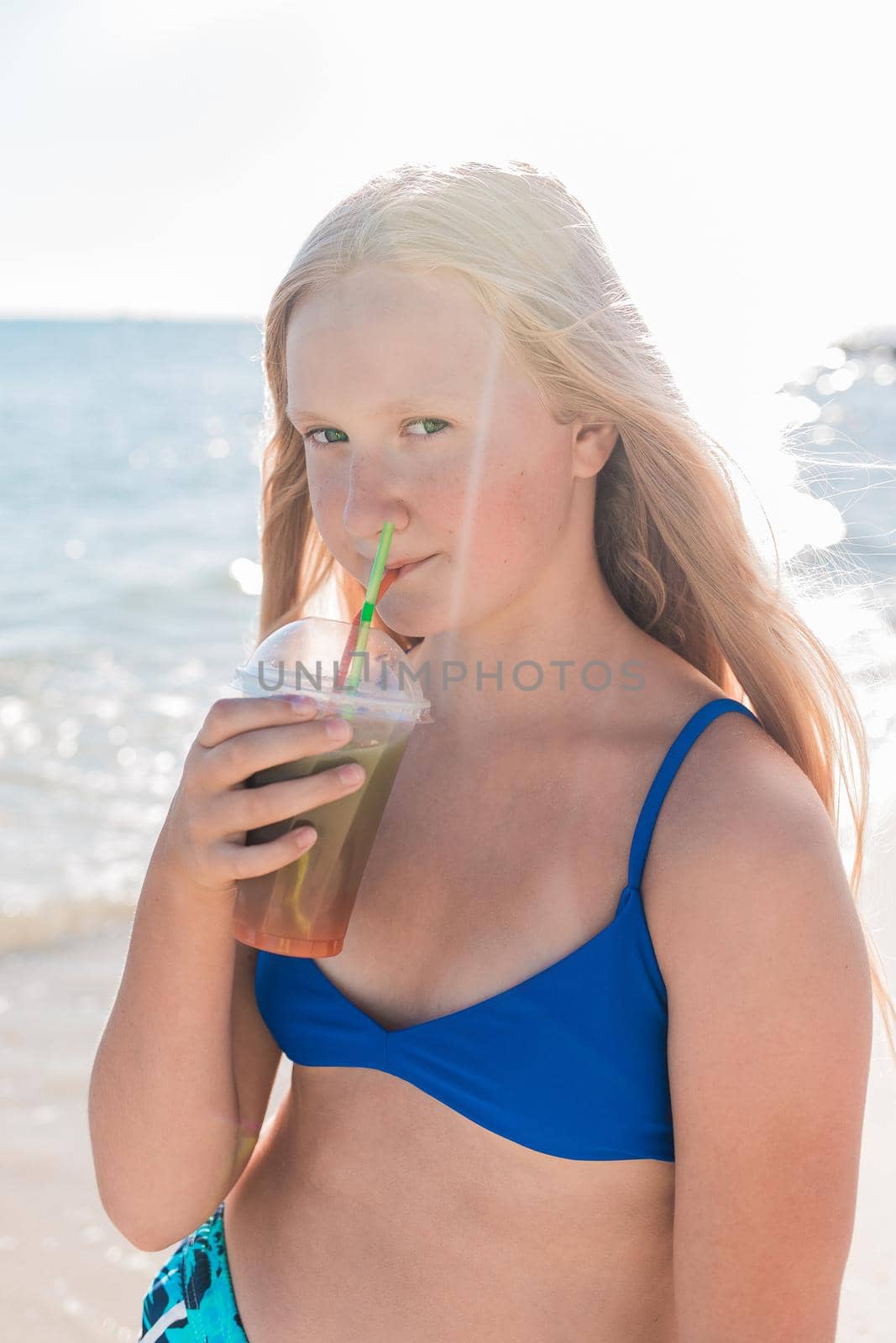 A young girl with blonde hair of European appearance, a teenager holds and drink a colored cold non-alcoholic cocktail in her hand against the background of the sea beach.