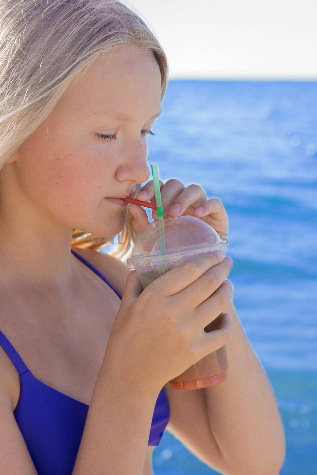 A young girl with blonde hair of European appearance, a teenager holds and drink a colored cold non-alcoholic cocktail in her hand against the background of the sea beach.