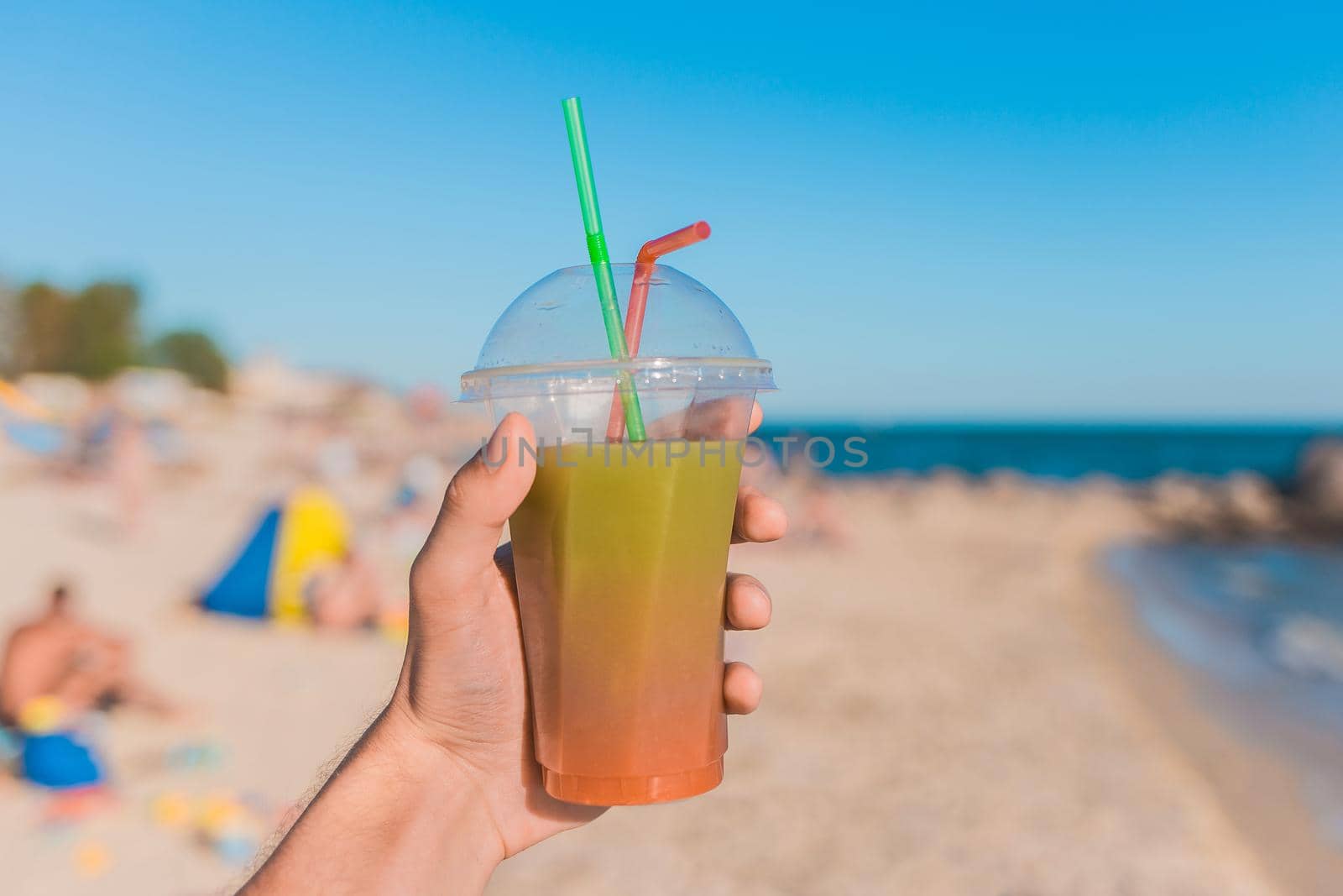 The guy's hand holds a colored chilled non-alcoholic cocktail against the background of the sea beach by AYDO8