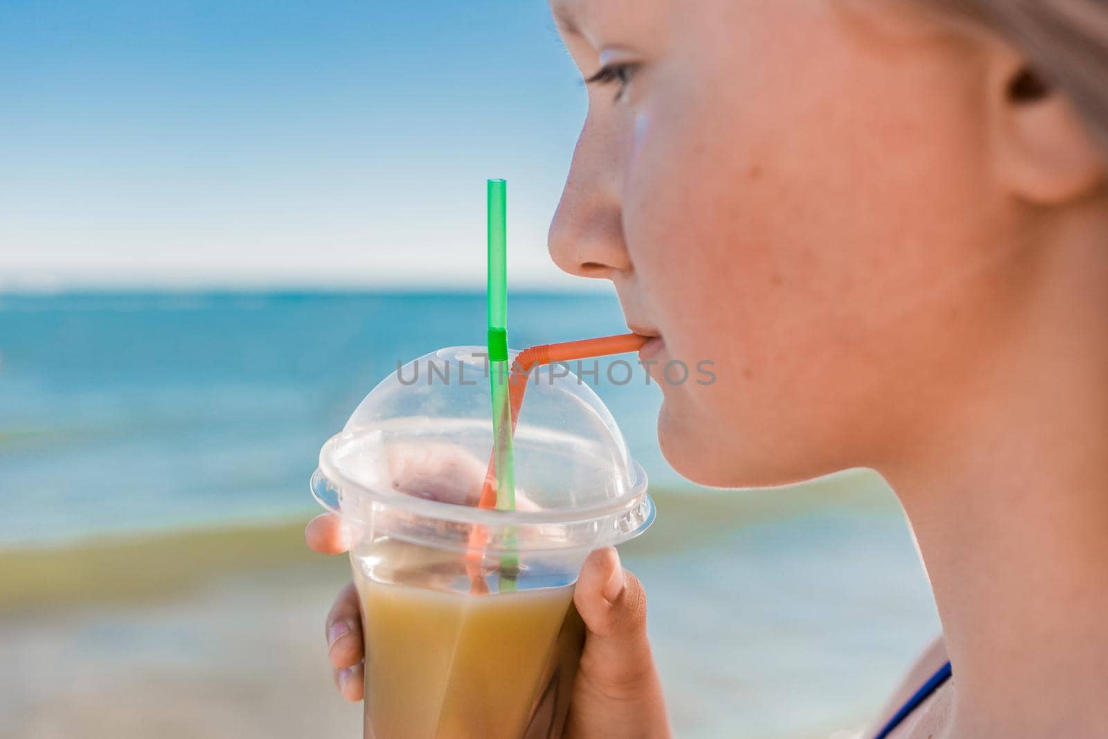 A young girl with blonde hair of European appearance, a teenager holds and drink a colored cold non-alcoholic cocktail in her hand against the background of the sea beach by AYDO8