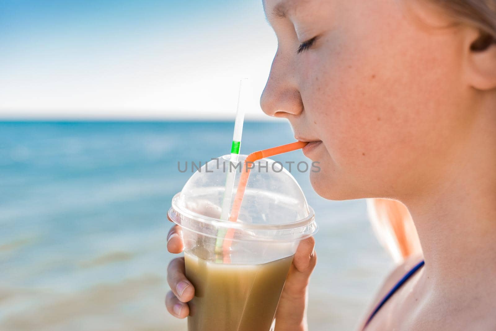 A young girl with blonde hair of European appearance, a teenager holds and drink a colored cold non-alcoholic cocktail in her hand against the background of the sea beach by AYDO8