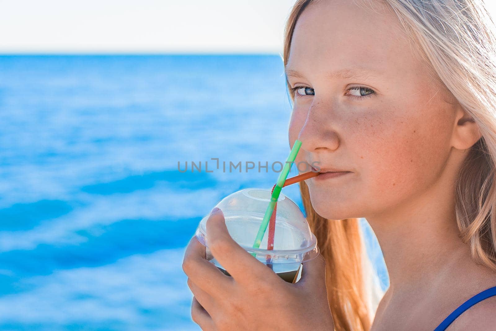 A young girl with blonde hair of European appearance, a teenager holds and drink a colored cold non-alcoholic cocktail in her hand against the background of the sea beach.