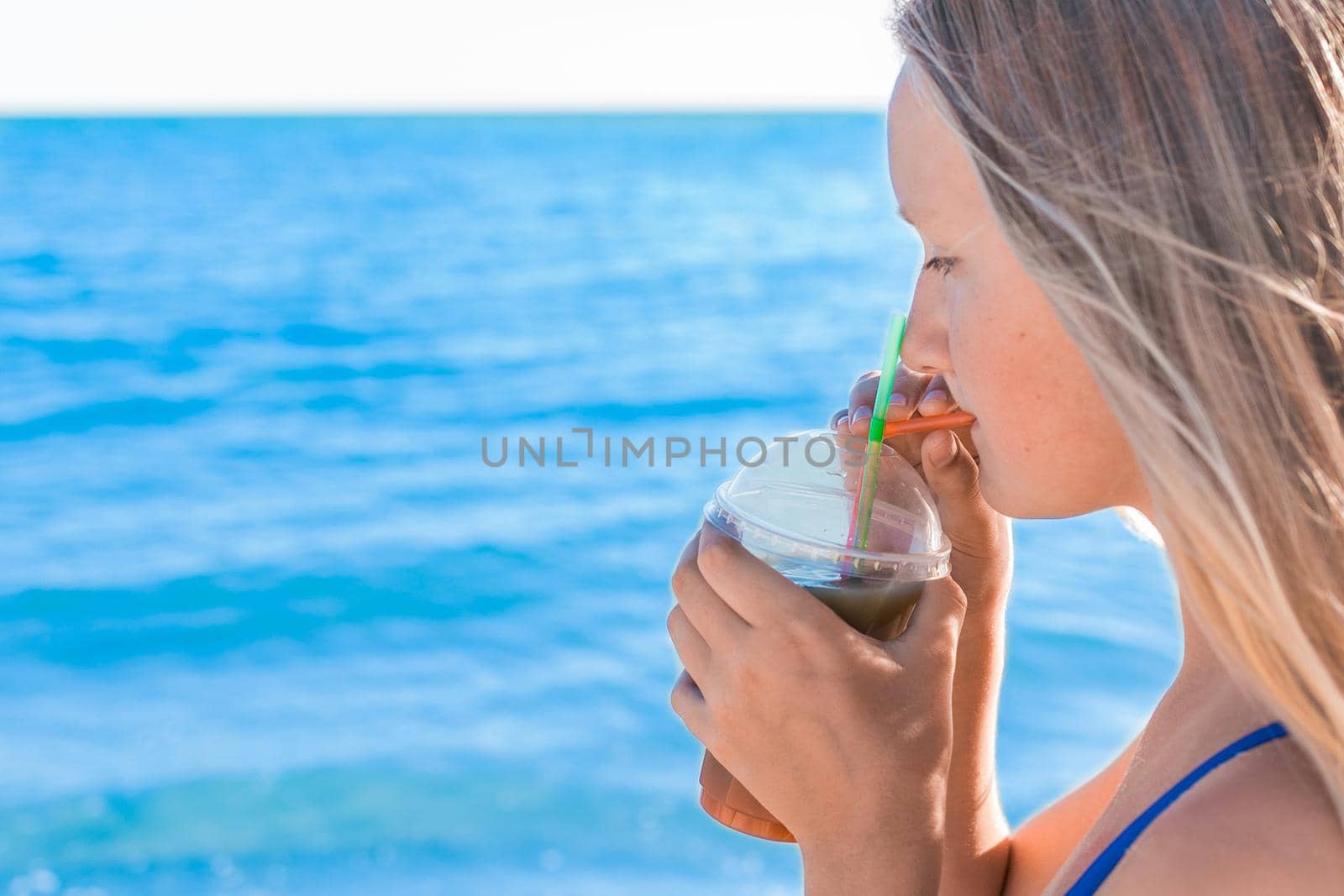 A young girl with blonde hair of European appearance, a teenager holds and drink a colored cold non-alcoholic cocktail in her hand against the background of the sea beach by AYDO8