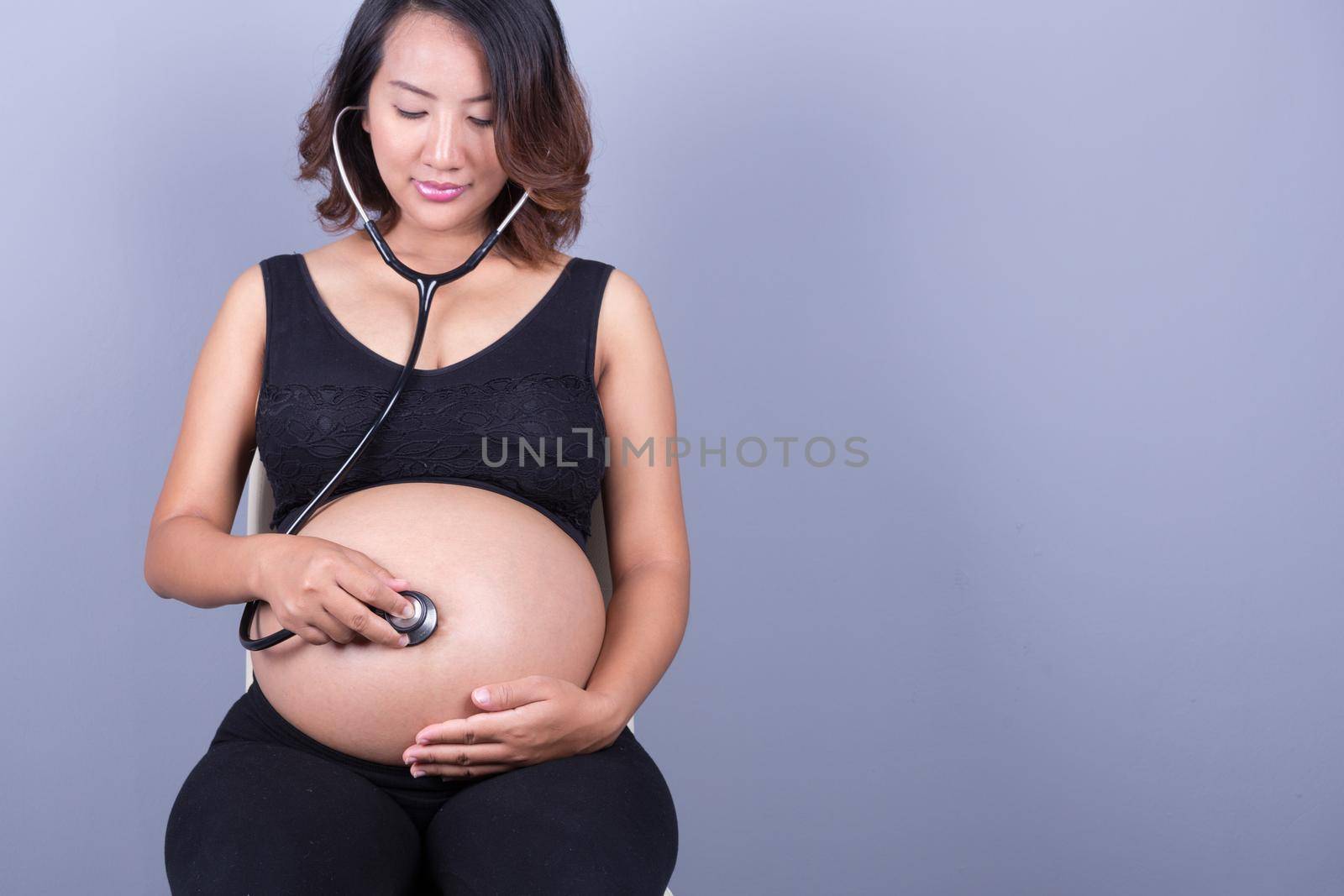 young pregnant asian woman with stethoscope listening belly to baby by geargodz