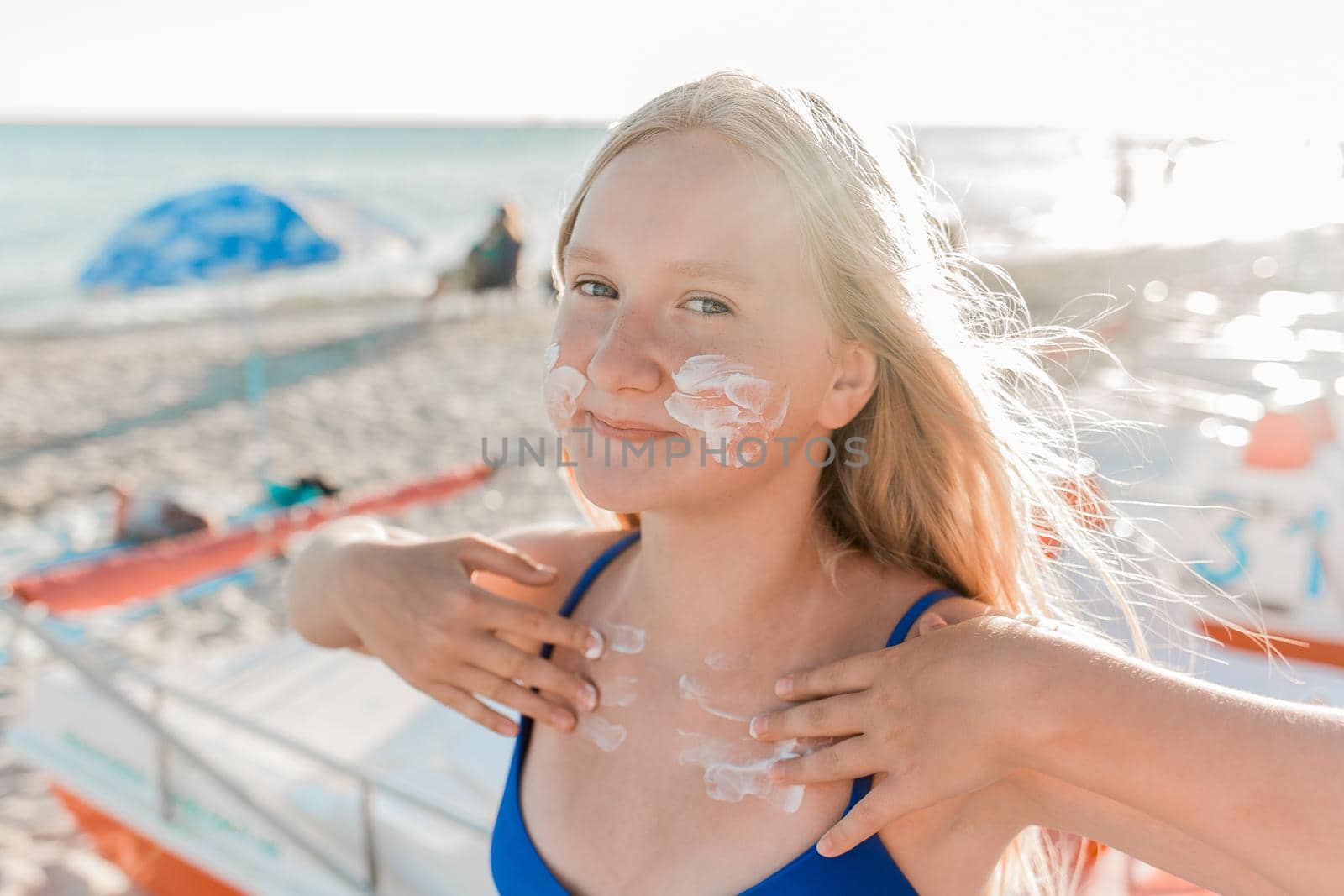 Portrait of a young positive teenage girl blonde of European appearance with sunscreen on her face and body against the background of a sea beach.