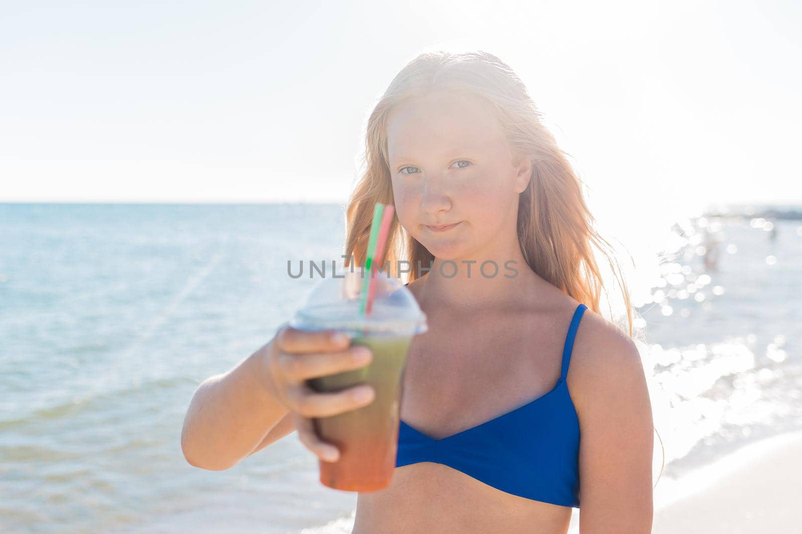 A young girl with blonde hair of European appearance, a teenager holds a colored cold non-alcoholic cocktail in her hand against the background of the sea beach.