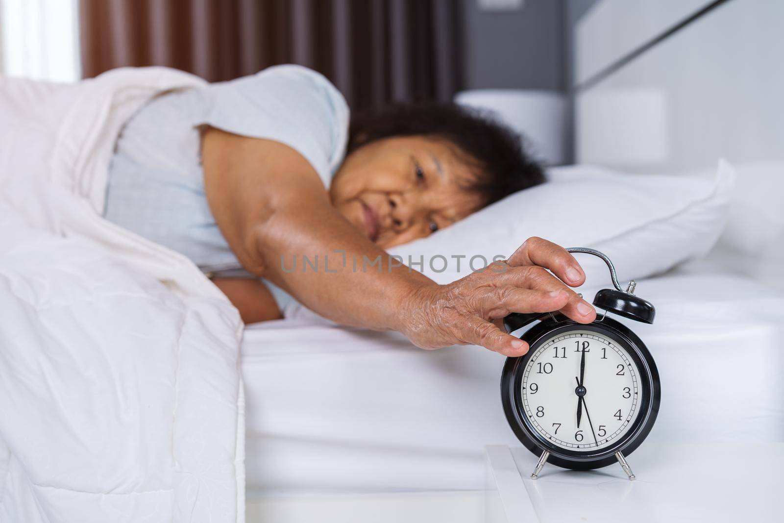 senior woman in bed pressing snooze button on morning alarm clock  by geargodz