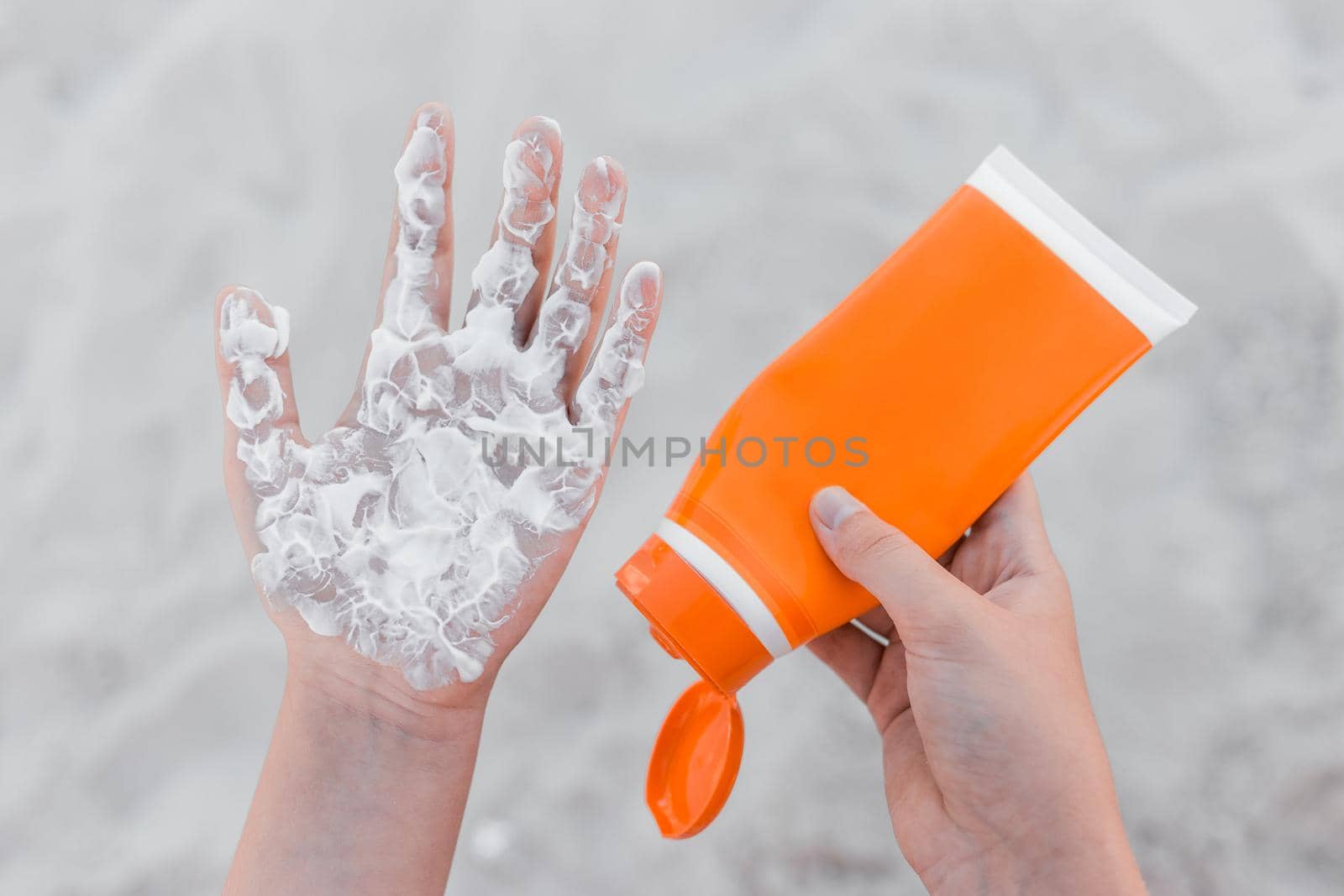 The girl holds sunscreen, in her hand and squeezes it on her arm palm to protect her from the sun against the background of beach sand.