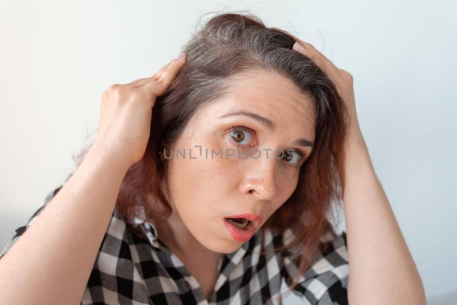 Horrified young woman looking in the bathroom mirror staring open mouthed at the first grey hair on her scalp, a first sign of ageing, or noticing that she is suffering from dandruff