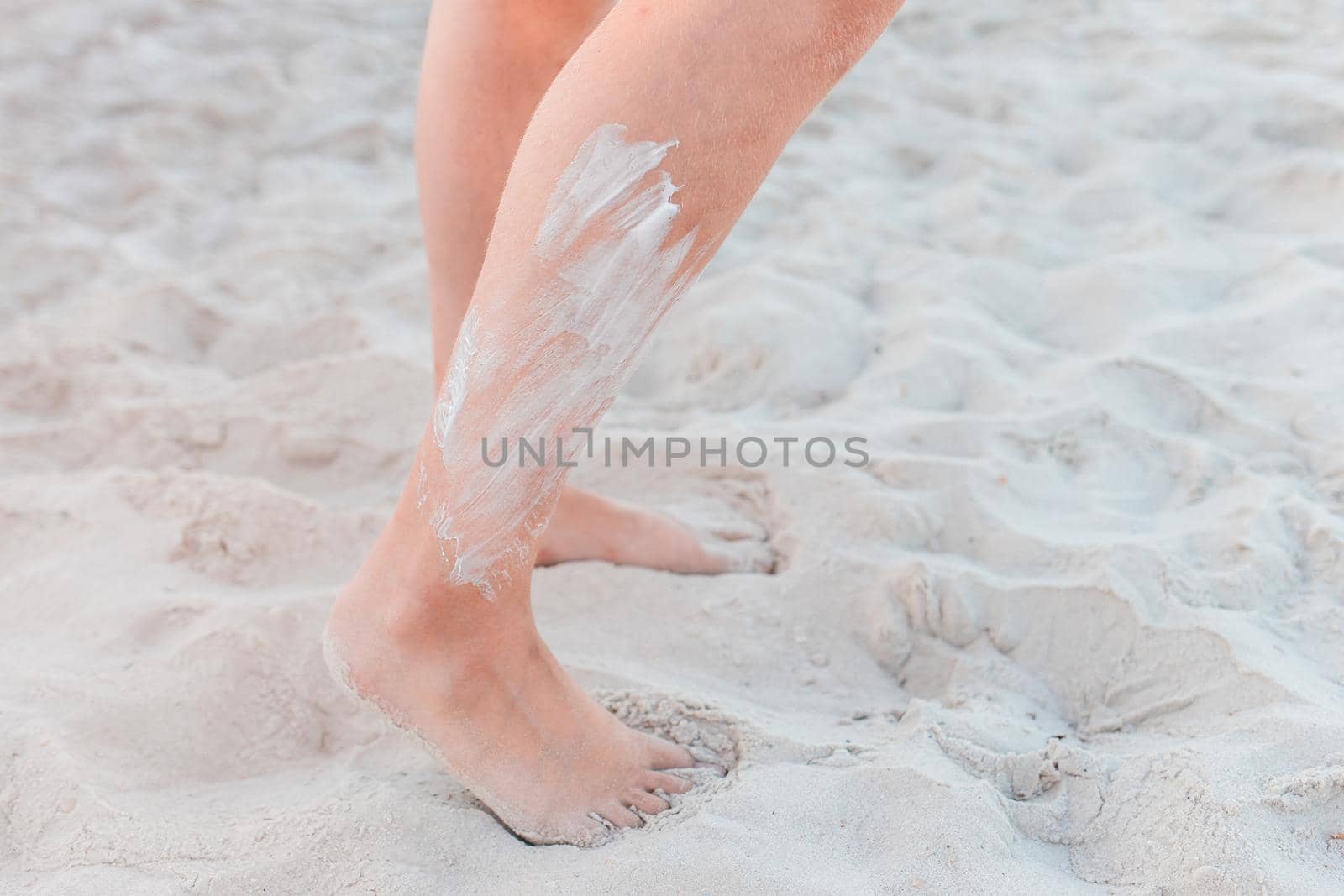 The legs of a young girl stand on the beach white sand with sunscreen on the skin by AYDO8