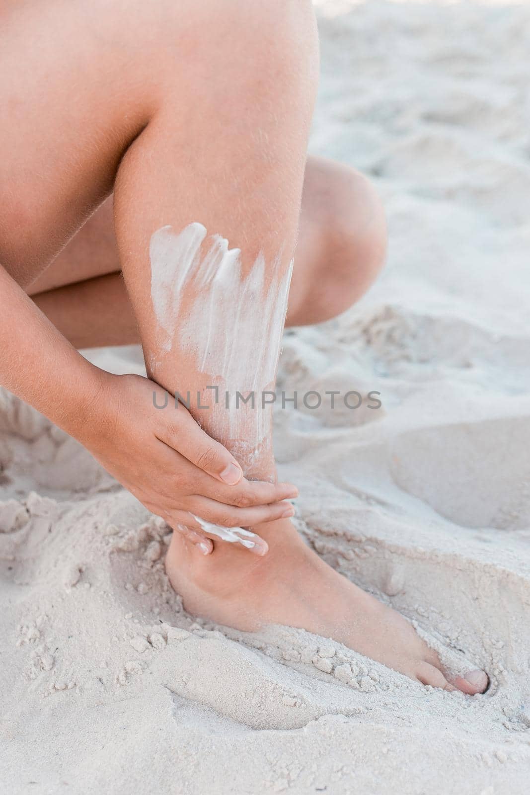 A young girl's hand smears sunscreen on her leg next to the beach sand.