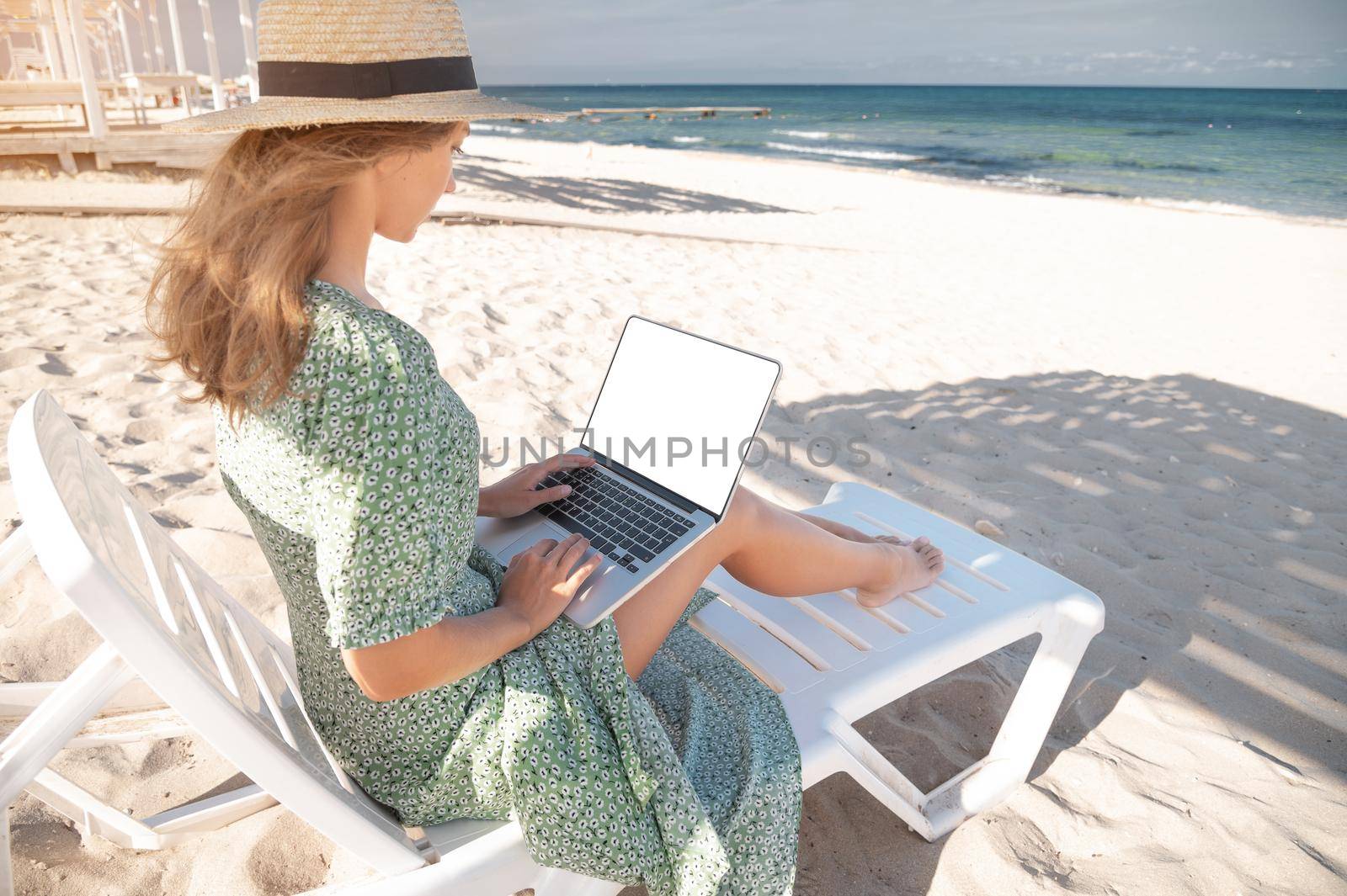 Rear view of an attractive young Caucasian woman in a green dress and hat sits on a lounger with a laptop in her hands in the shade of a wooden umbrella. Cut out laptop screen by yanik88