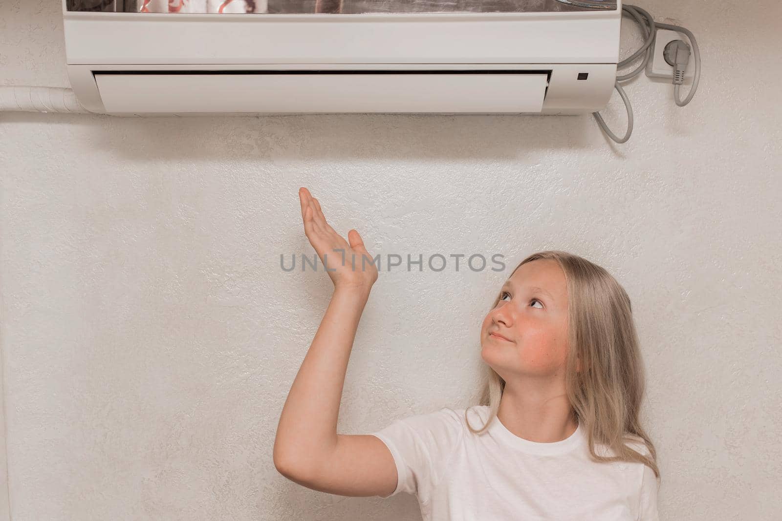 Young cute girl teenager blonde European appearance brings his hand to the air conditioner on the wall to check the heat and cold, the temperature in the room.