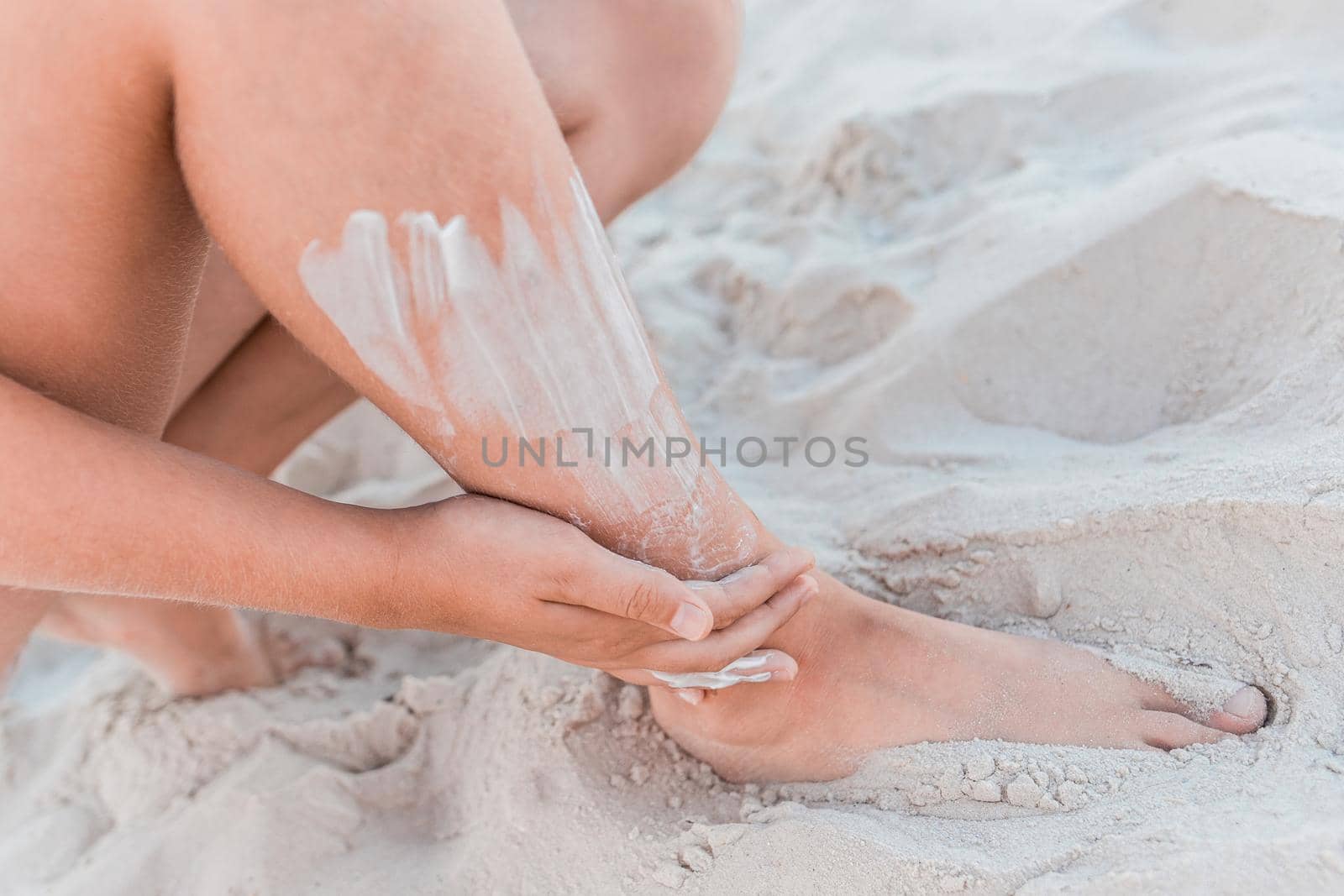 A young girl's hand smears sunscreen on her leg next to the beach sand.