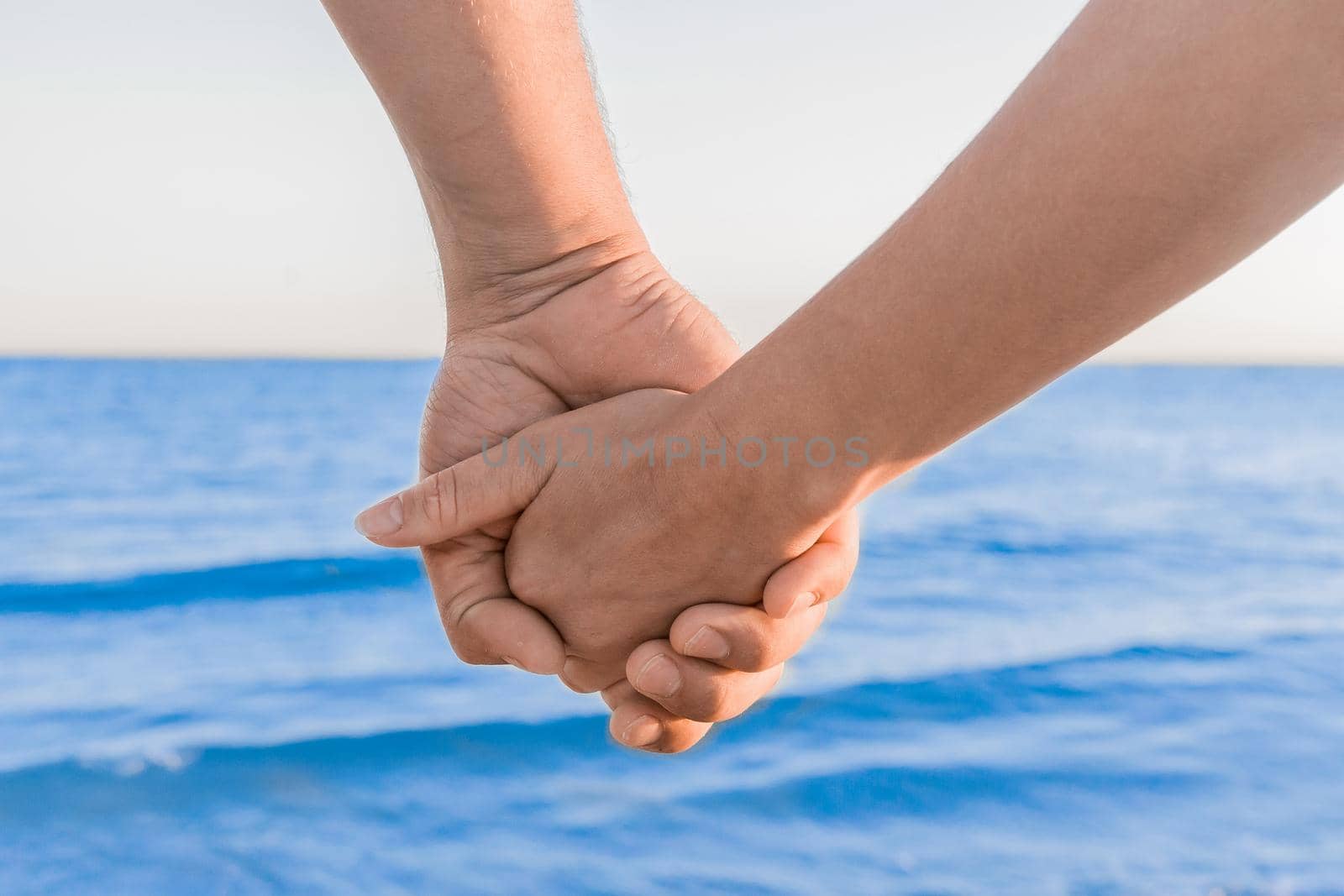 The hands of a man and a woman are held together against the background of the sea, a couple in love and romance on vacation by AYDO8