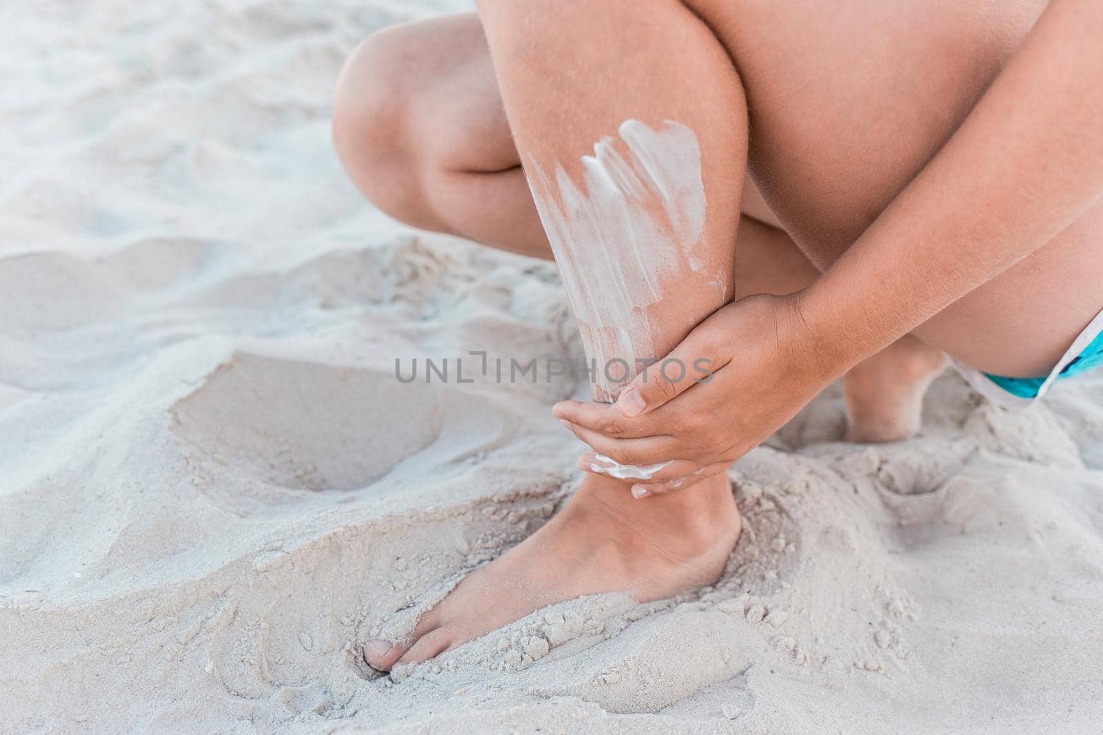 A young girl's hand smears sunscreen on her leg next to the beach sand.