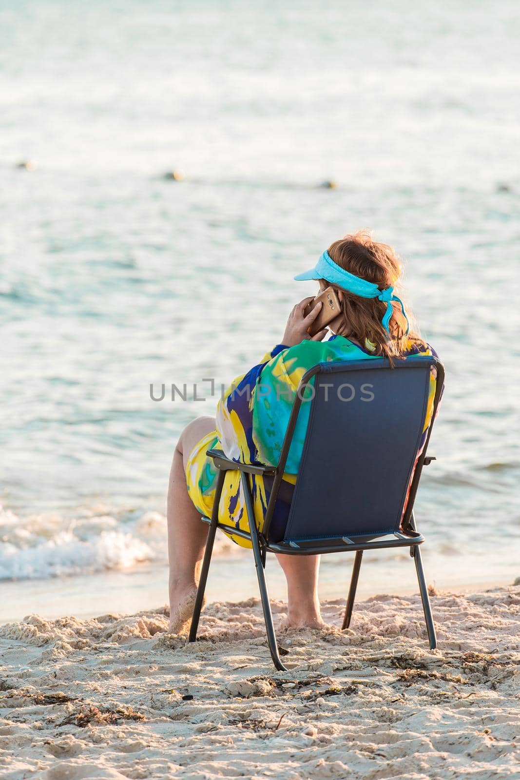 An adult woman in a pareo sits on a folding chair by the sea water on the shore, and talks on a smartphone or mobile phone by AYDO8